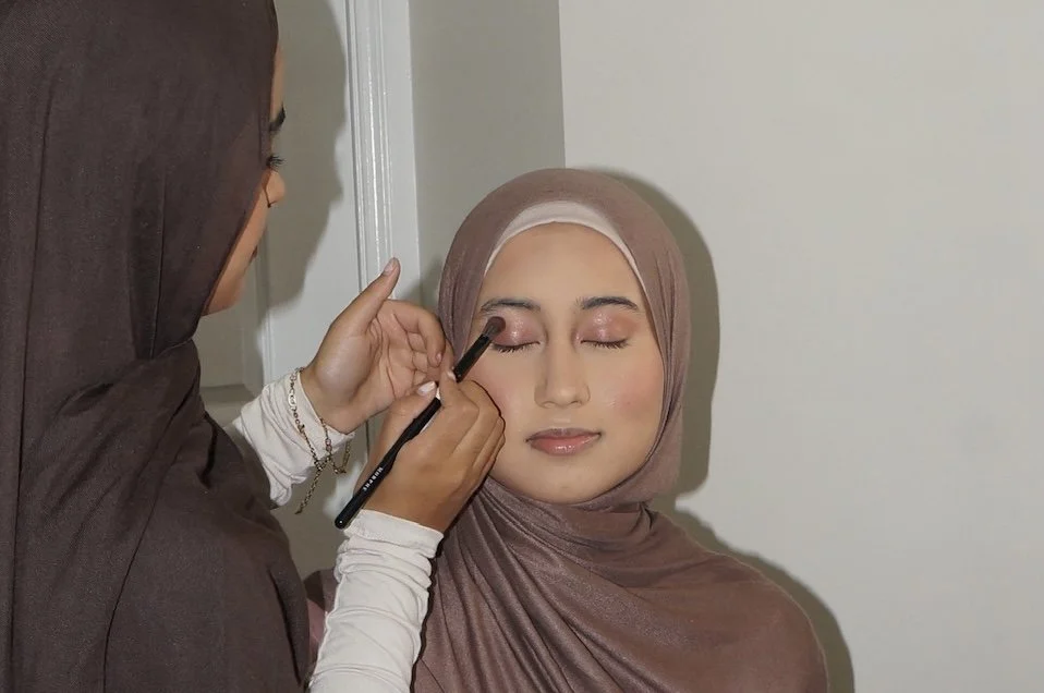 A makeup artist applying eyeshadow to a woman with a headscarf, who has her eyes closed.