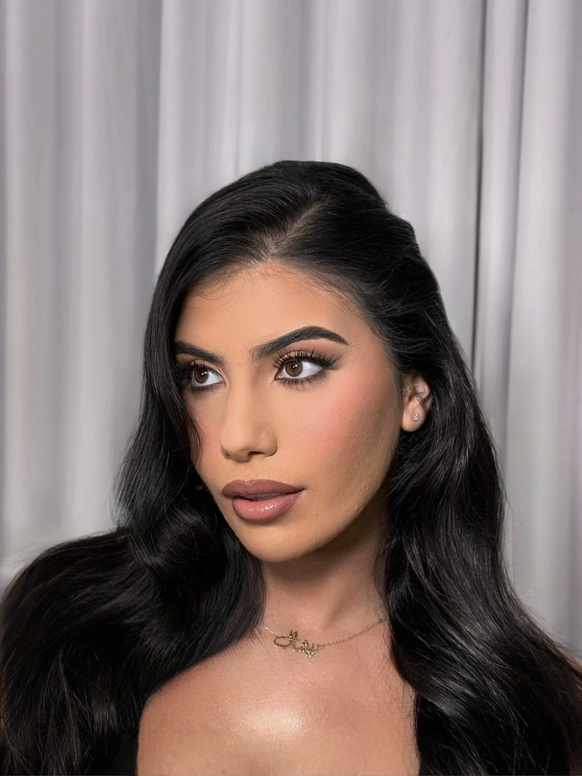 Portrait of a woman with long, dark, wavy hair looking to her right, wearing makeup and a small necklace, with a white curtain background.