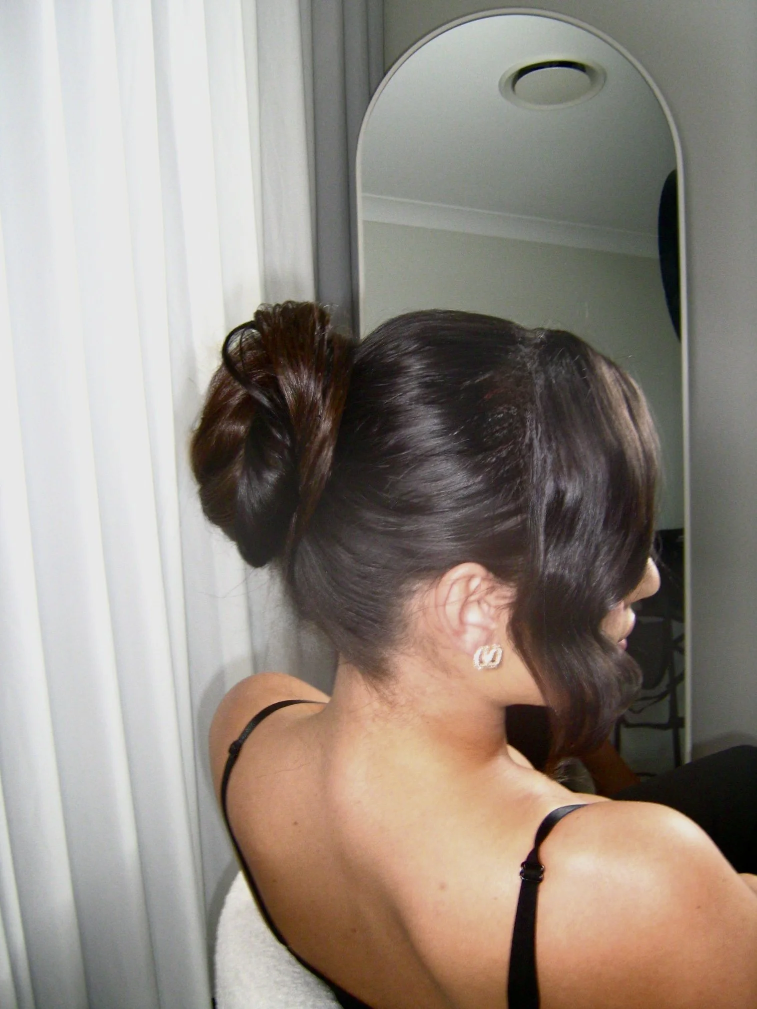 Woman with dark, styled hair in an elegant updo, wearing earrings, sitting in front of a mirror.