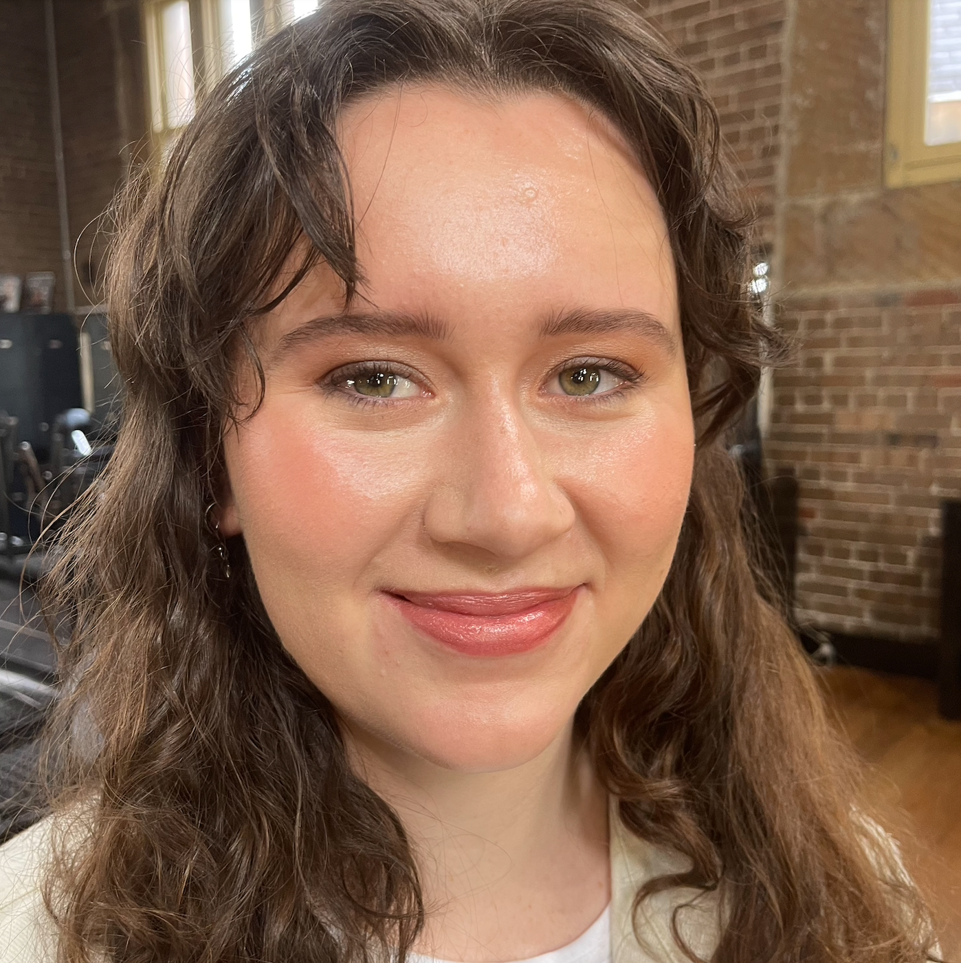 Close-up of a woman with wavy brown hair, wearing makeup with natural tones, smiling in an indoor space with brick walls and a wooden window frame.
