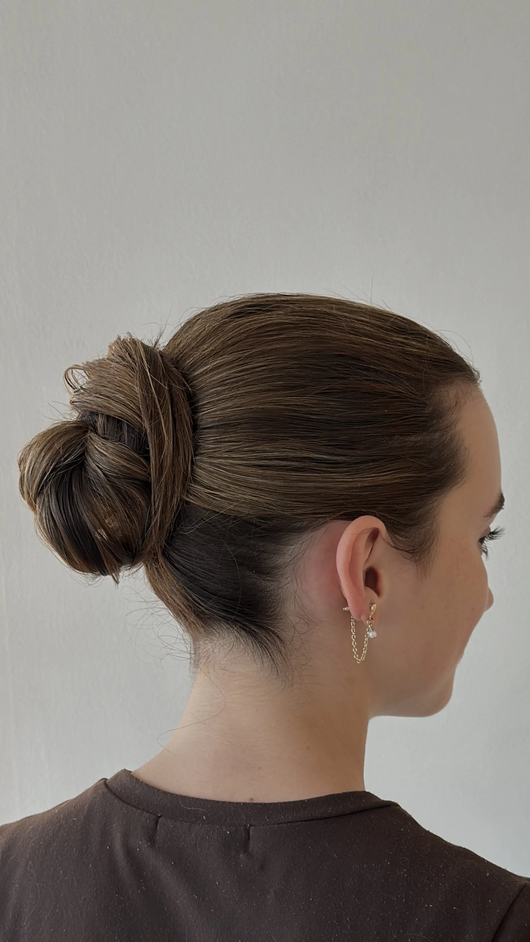 A woman with brown hair styled in a bun, wearing earrings, and a dark-colored top, standing against a plain white wall.
