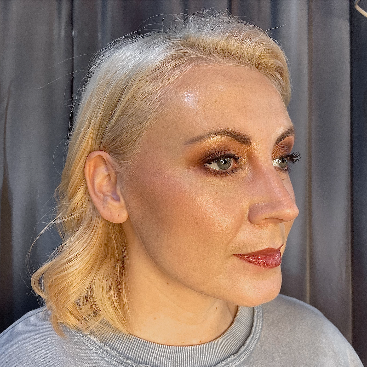 Close-up of a woman with blonde hair and makeup, looking to the right, wearing a light gray top, with a dark background.