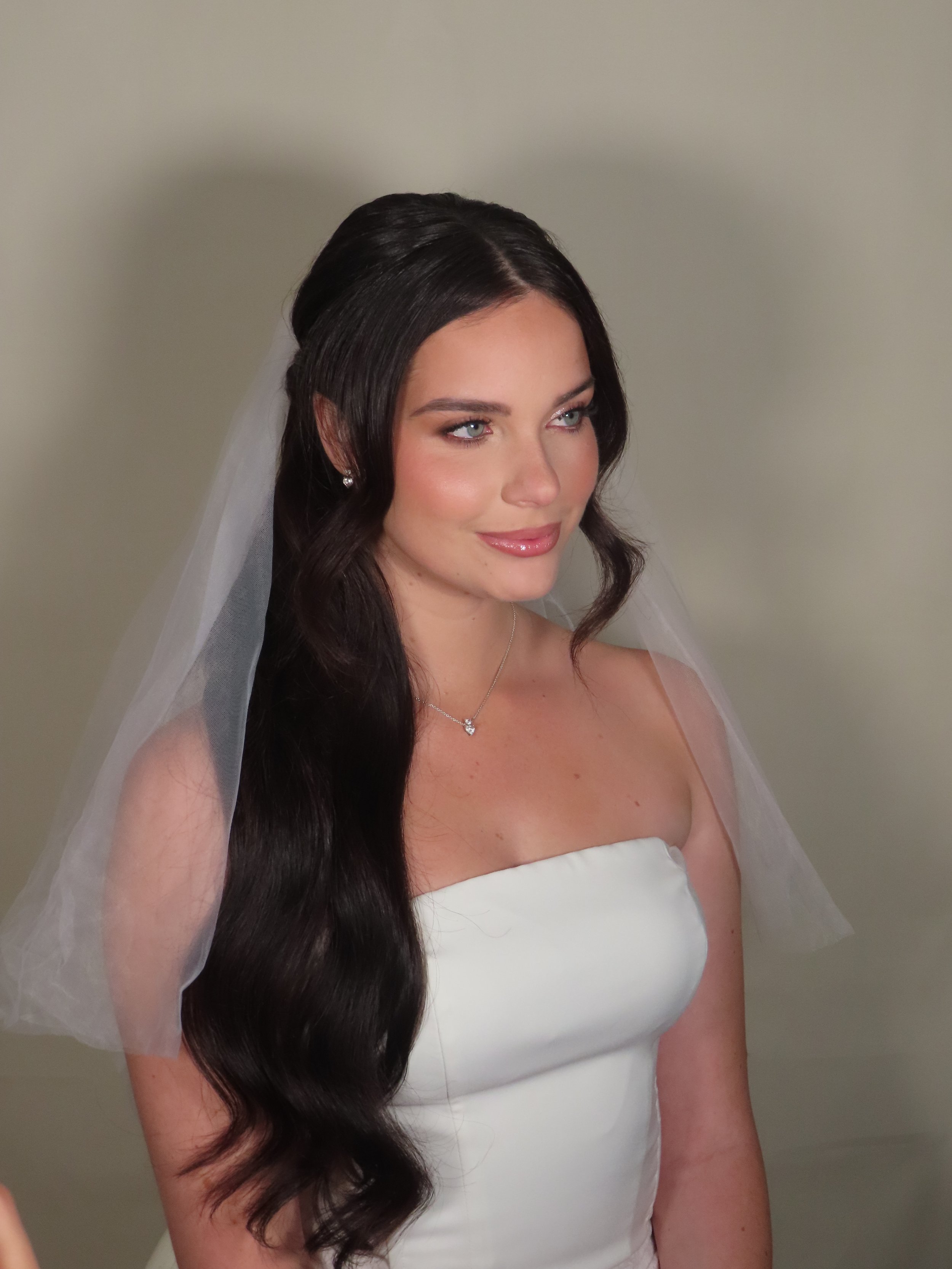 A young woman in a white strapless wedding dress with long dark wavy hair and a sheer veil, smiling softly, wearing earrings and a necklace, against a plain beige background. Half up half down wedding bridal hairstyling.  Soft glam natural makeup.