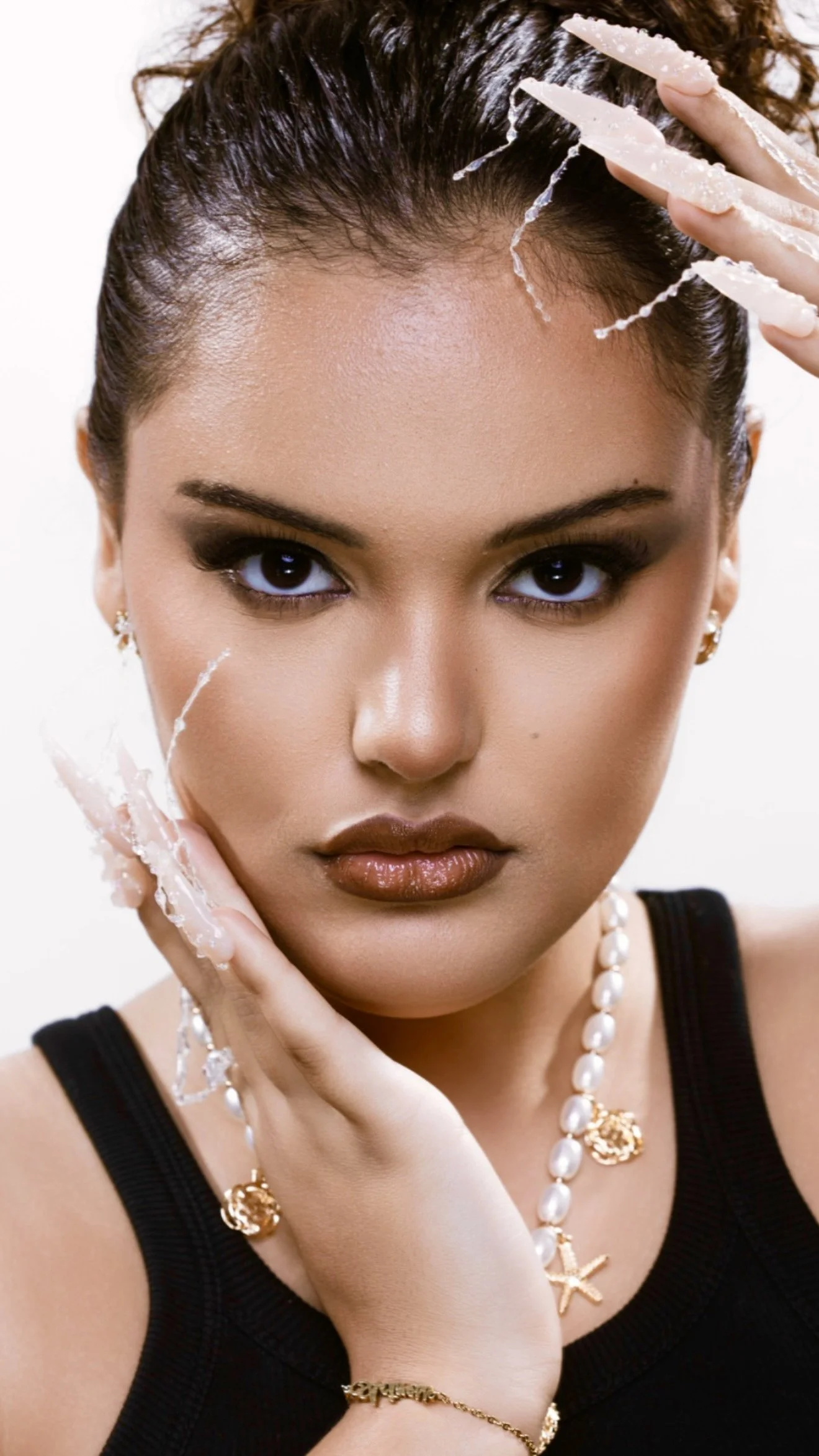 A woman with dark hair and brown eyes posing with water or a similar liquid on her face and fingers, wearing jewelry including a pearl necklace, starfish jewelry, and earrings, dressed in a black top.