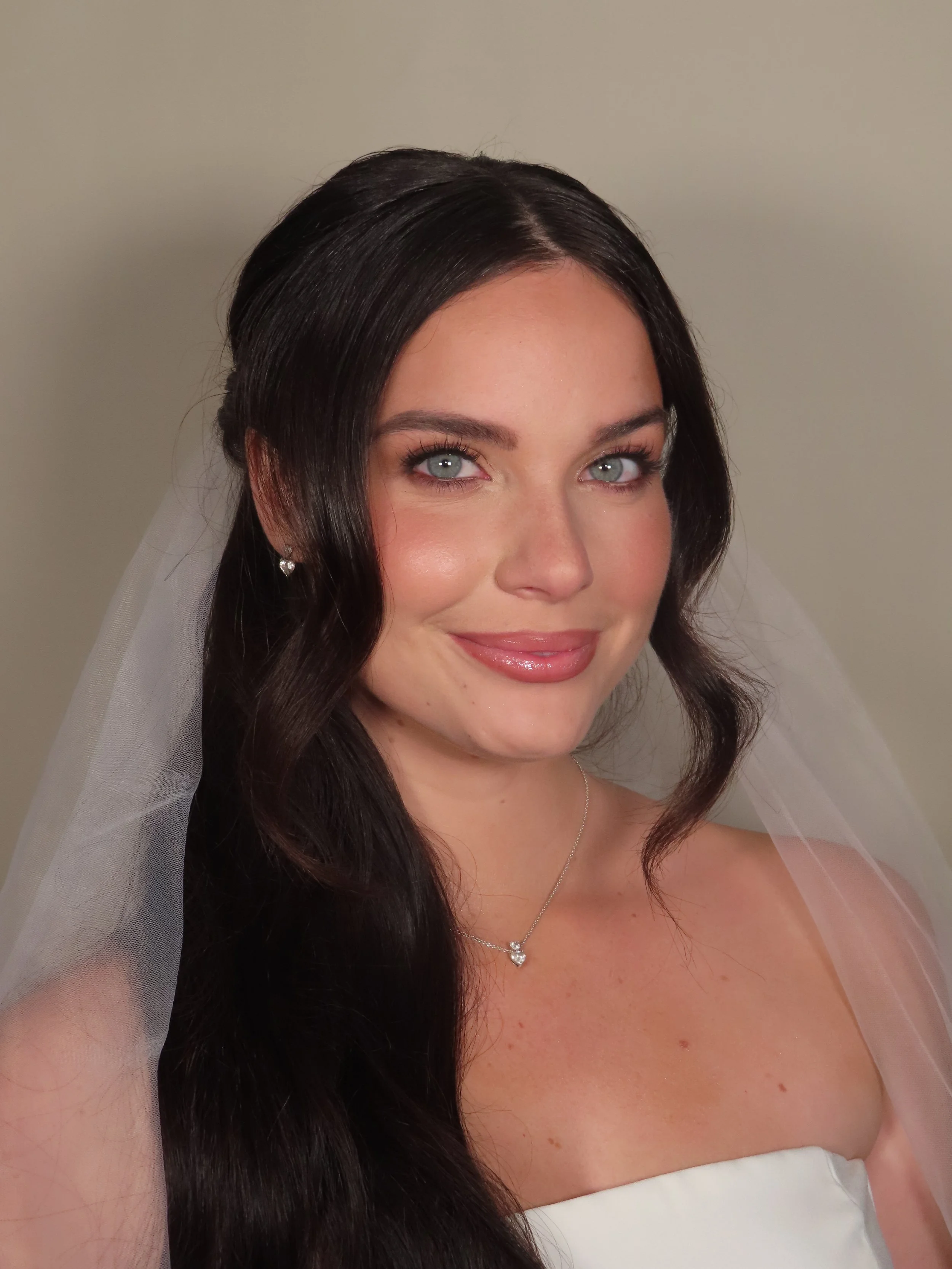 Portrait of a bride with long dark hair, blue eyes, wearing a white dress, veil, and jewelry, smiling softly.