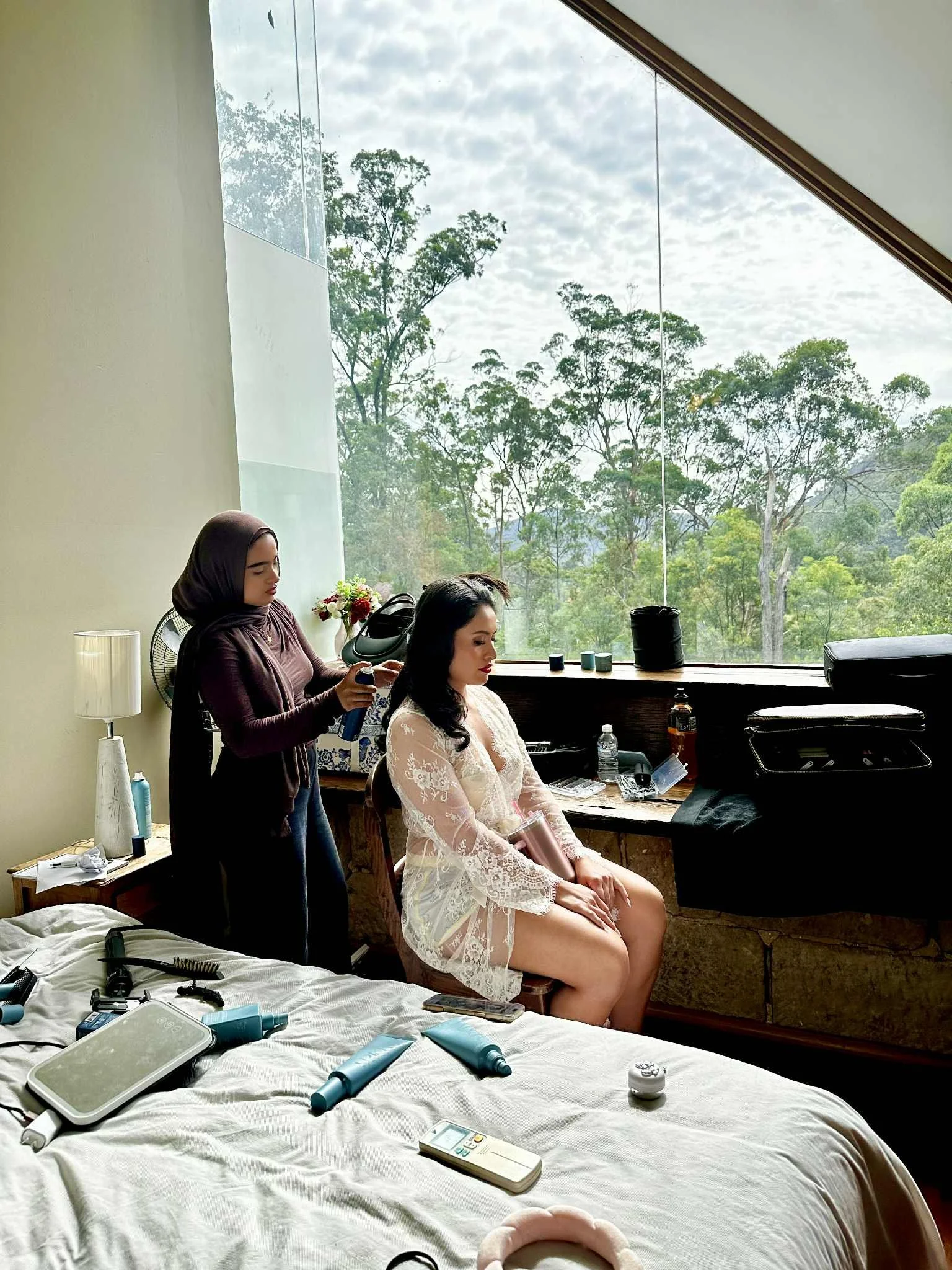 A woman in a white lace robe sitting on a chair in front of a large window with trees outside, getting her hair done by a stylist in a dark hijab. The room has a bed with hairstyling tools and products on it.