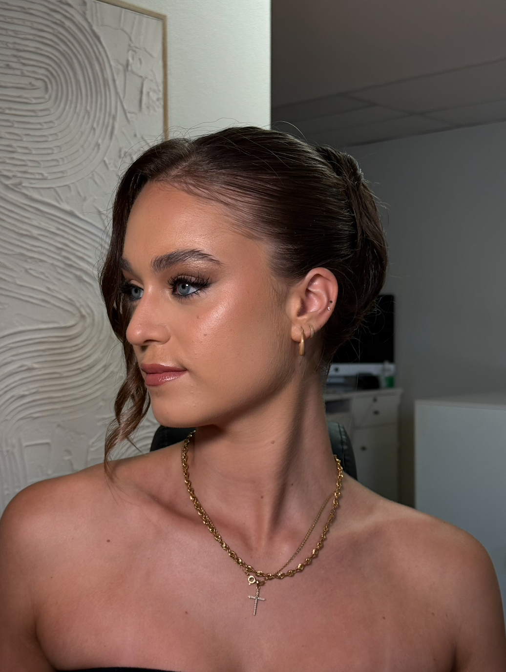A woman with elegant makeup and hairstyle, wearing gold jewelry including layered necklaces with a cross pendant, and small earrings, in an indoor setting with textured wall art in the background.