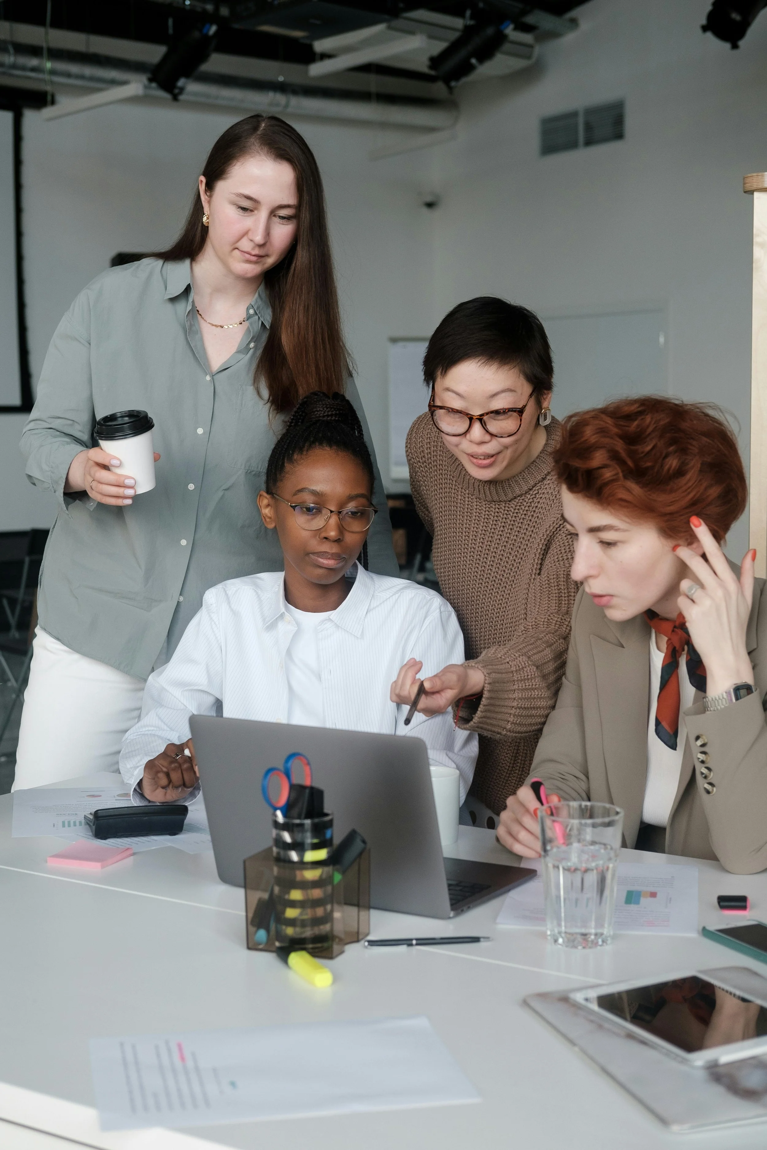 Five women are gathered around a table in a modern office, working together on a project using a laptop and documents, with various office supplies and a glass of water on the table.