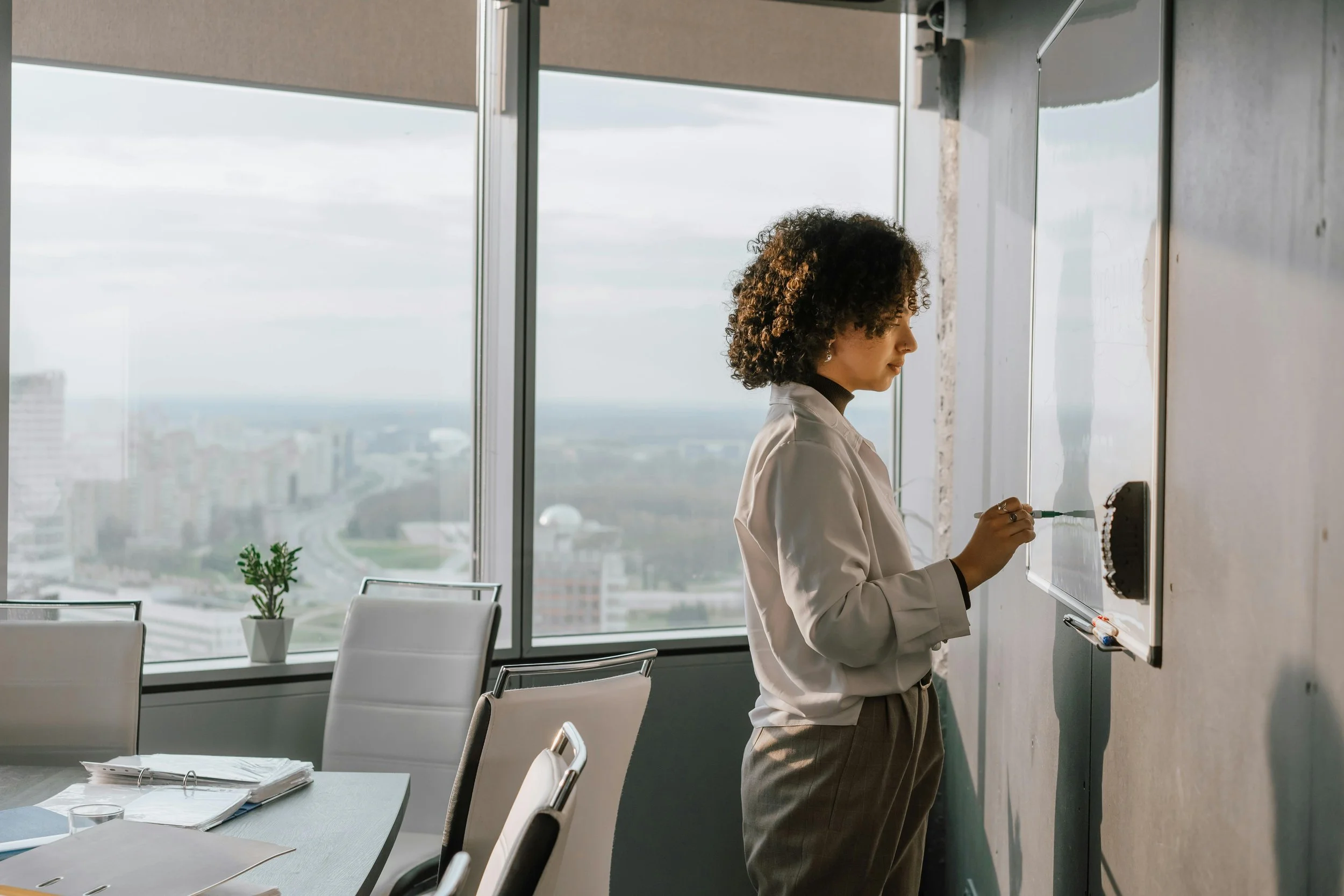 A woman in professional attire writing on a whiteboard in a modern office with large windows overlooking a cityscape.