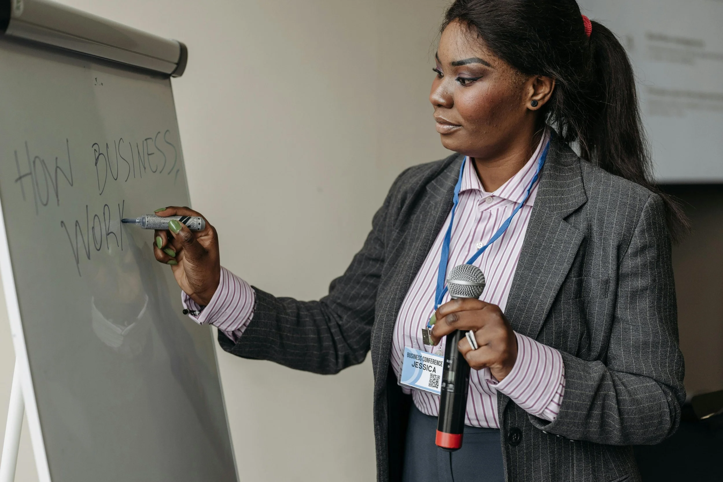 A woman in professional attire writing on a whiteboard with a marker. She is holding a microphone and has a conference badge around her neck.