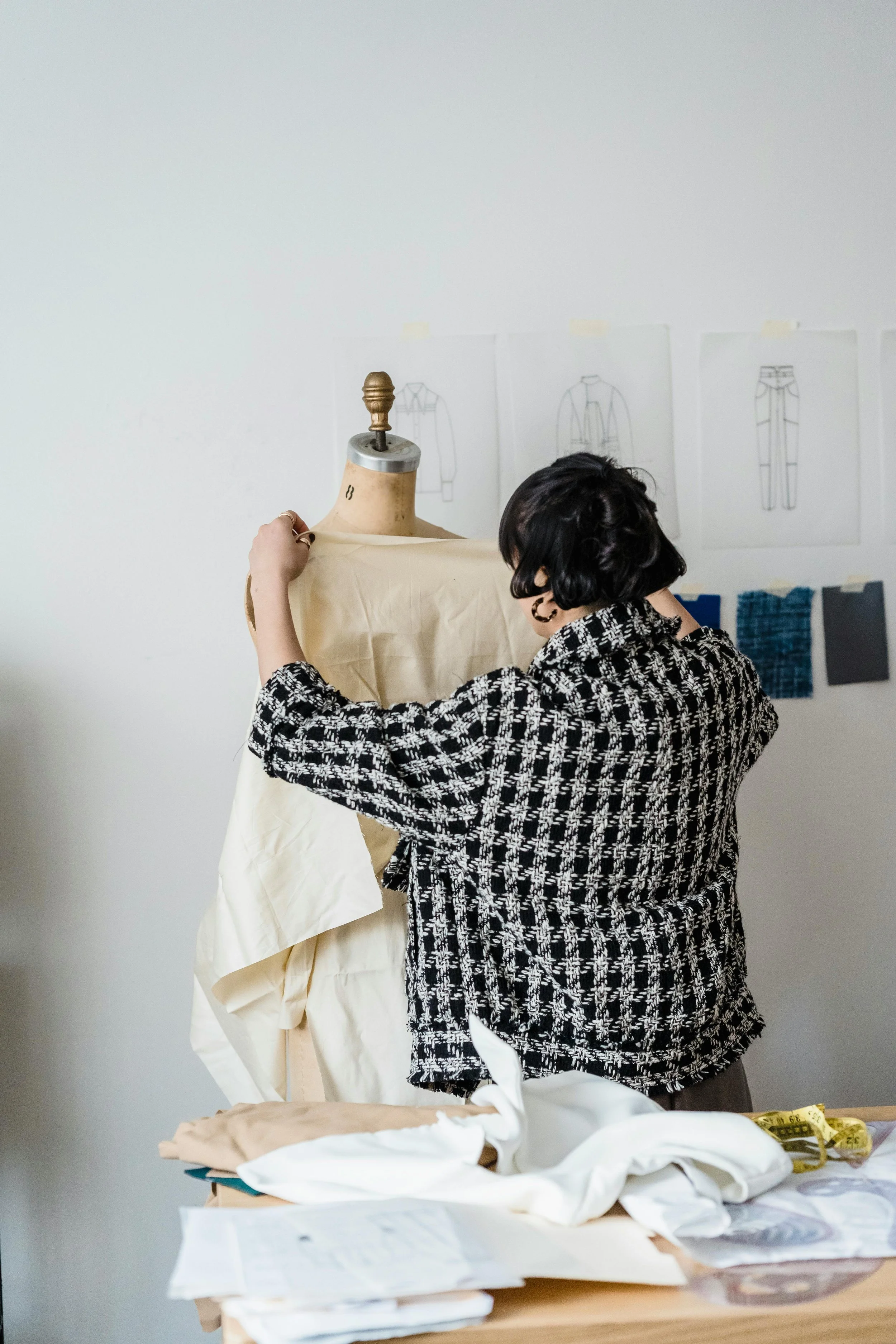 A person with short dark hair and hoop earrings, wearing a black and white patterned sweater, is adjusting a beige dress on a mannequin. The workspace includes fabric, pattern sketches taped to the wall, and sewing tools.
