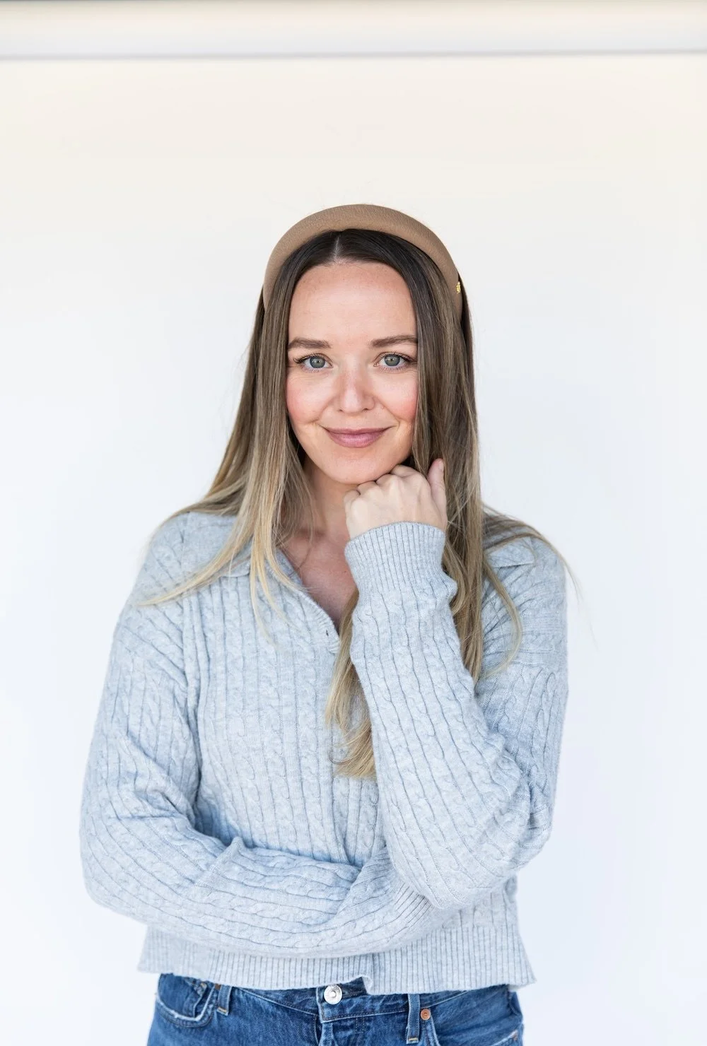 A young woman with long blonde hair wearing a beige headband and a light gray cable-knit sweater, smiling with her chin resting on her hand against a plain white background.