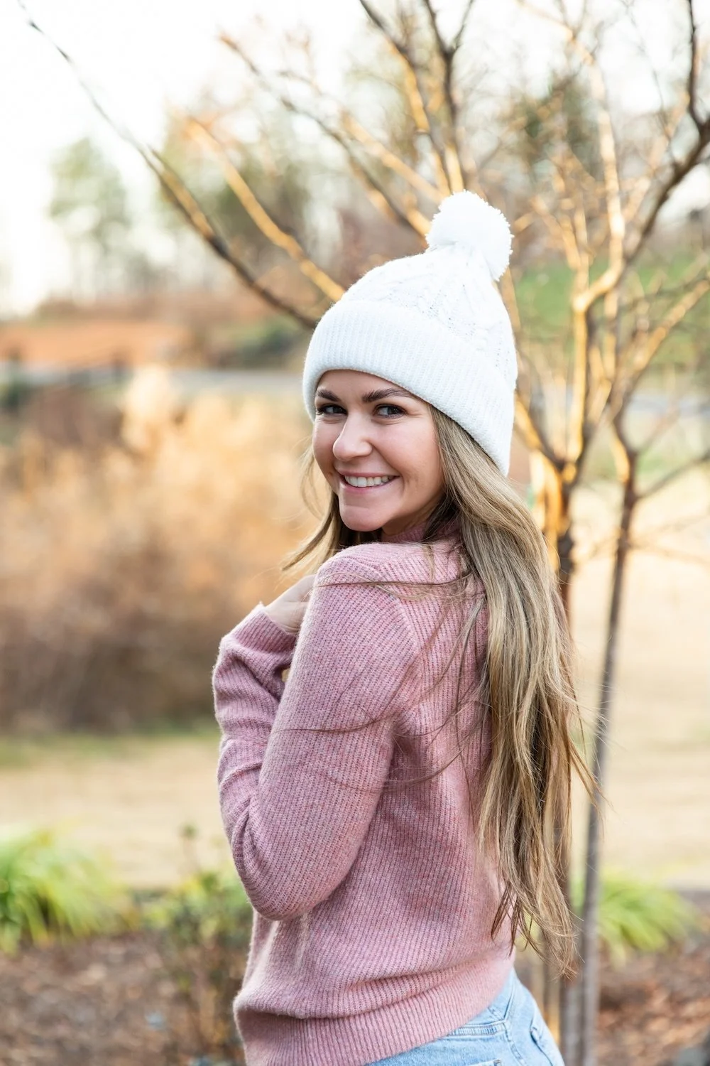 A woman smiling outdoors wearing a white knit hat with a pom-pom and a pink sweater.
