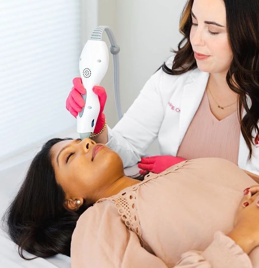 A healthcare professional performs a cosmetic procedure on a patient using a handheld device. The patient is lying down with eyes closed, and the professional wears pink gloves and a white coat in a clinical setting.
