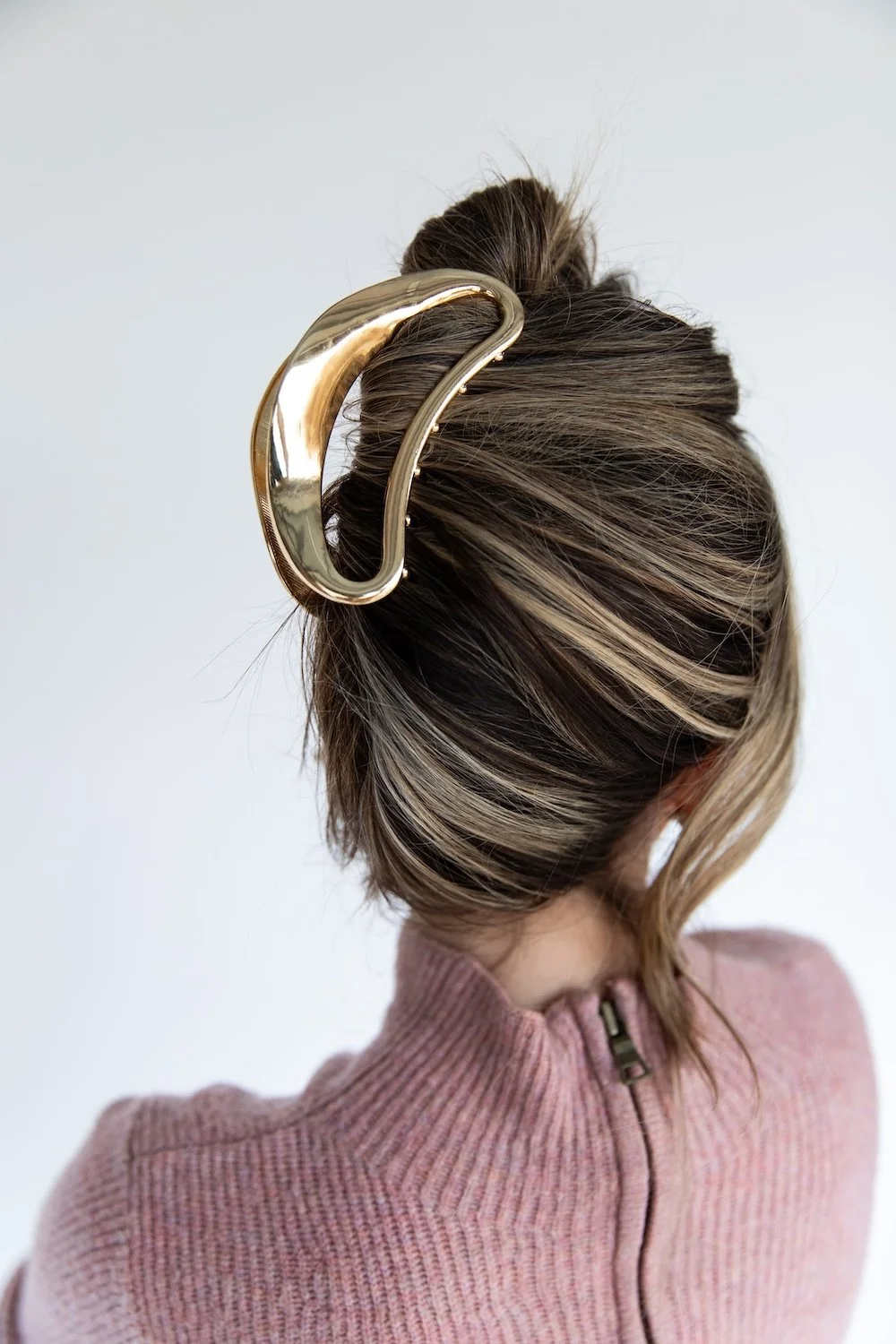 A woman with brown and blonde hair in a messy bun, secured with a large, shiny gold hair clip, wearing a pink sweater with a zipper at the back.