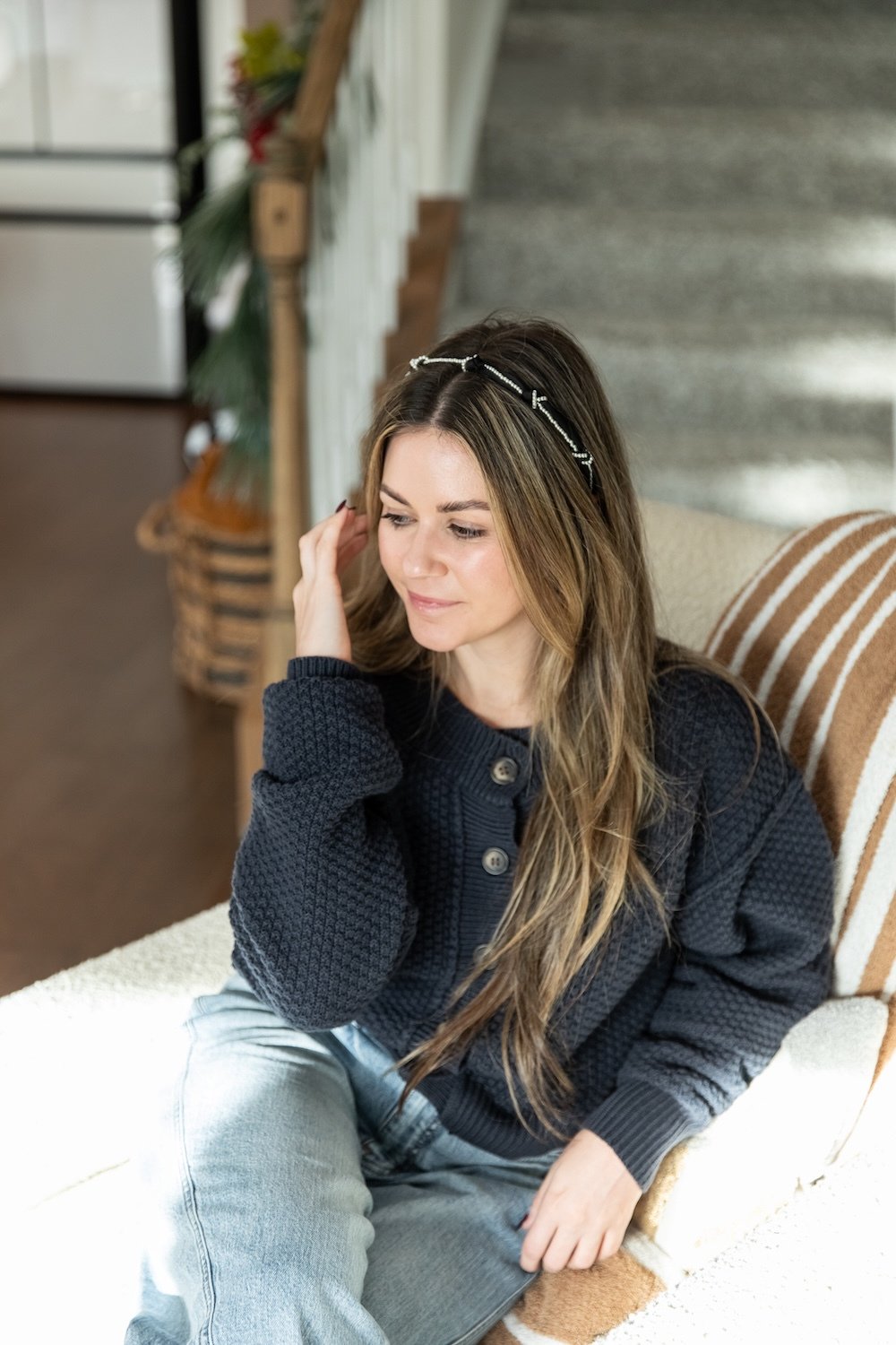 A woman with long, wavy brown hair wearing a black knit sweater and light blue jeans, sitting on a striped armchair in a cozy, decorated room, with a thoughtful expression.