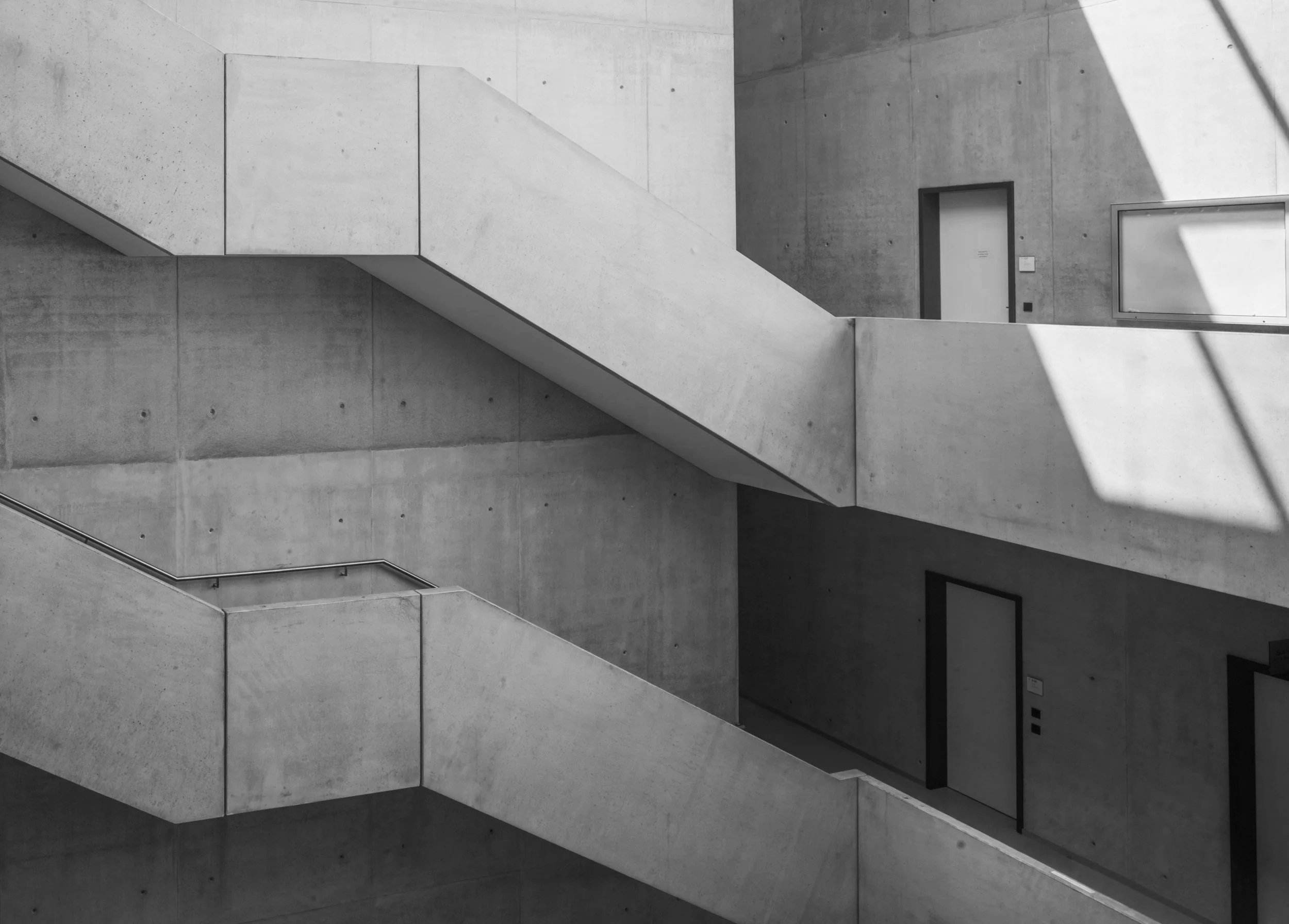 Black and white photo of a modern concrete building with stairs and doors, featuring shadows from sunlight.