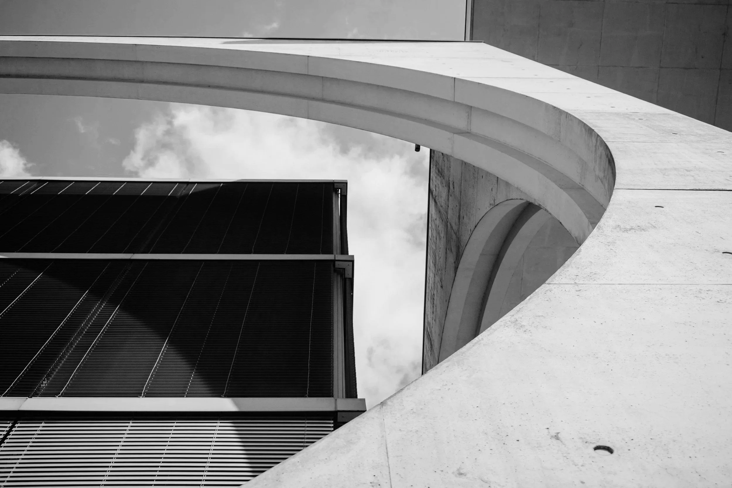 Black and white photograph of modern architectural structures, featuring a curved concrete arch and a building with vertical louvers, set against a background of cloudy sky.