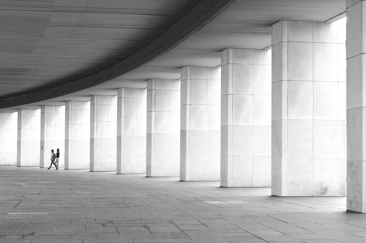 A black and white photograph of a large, empty building with tall rectangular columns supporting a curved ceiling. Two people are walking near the columns.