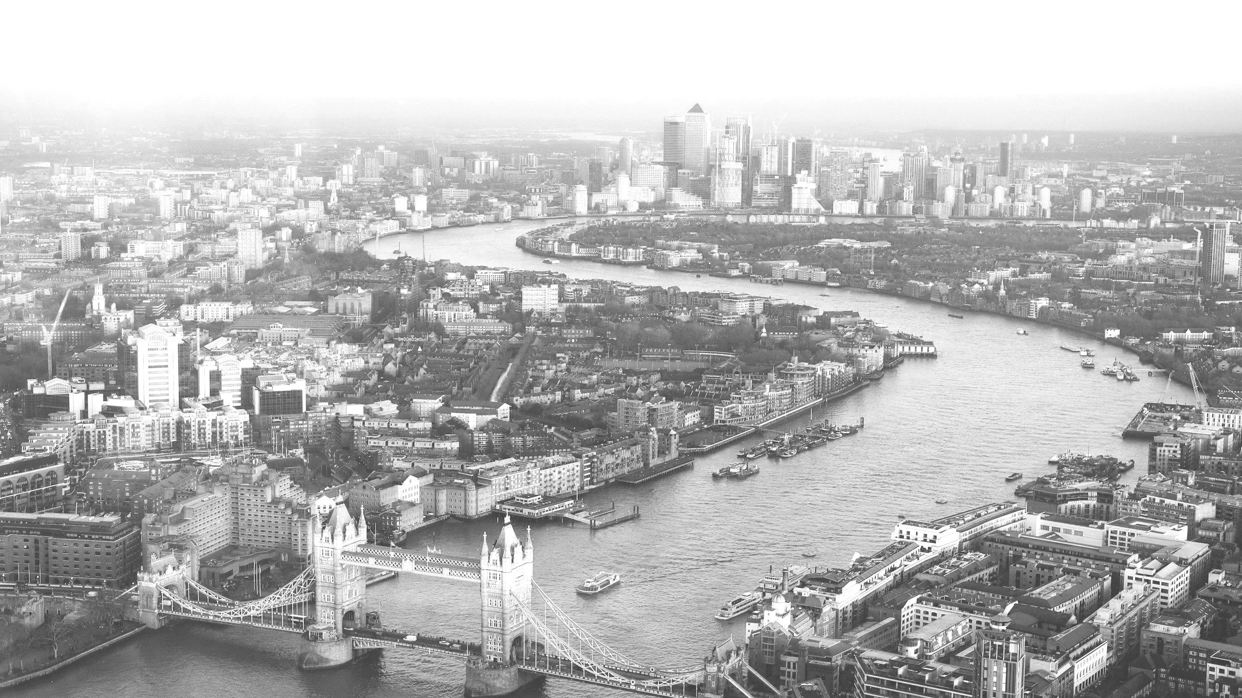 Black and white aerial view of London showing Tower Bridge, River Thames, and city skyline with numerous buildings and ships on the river.