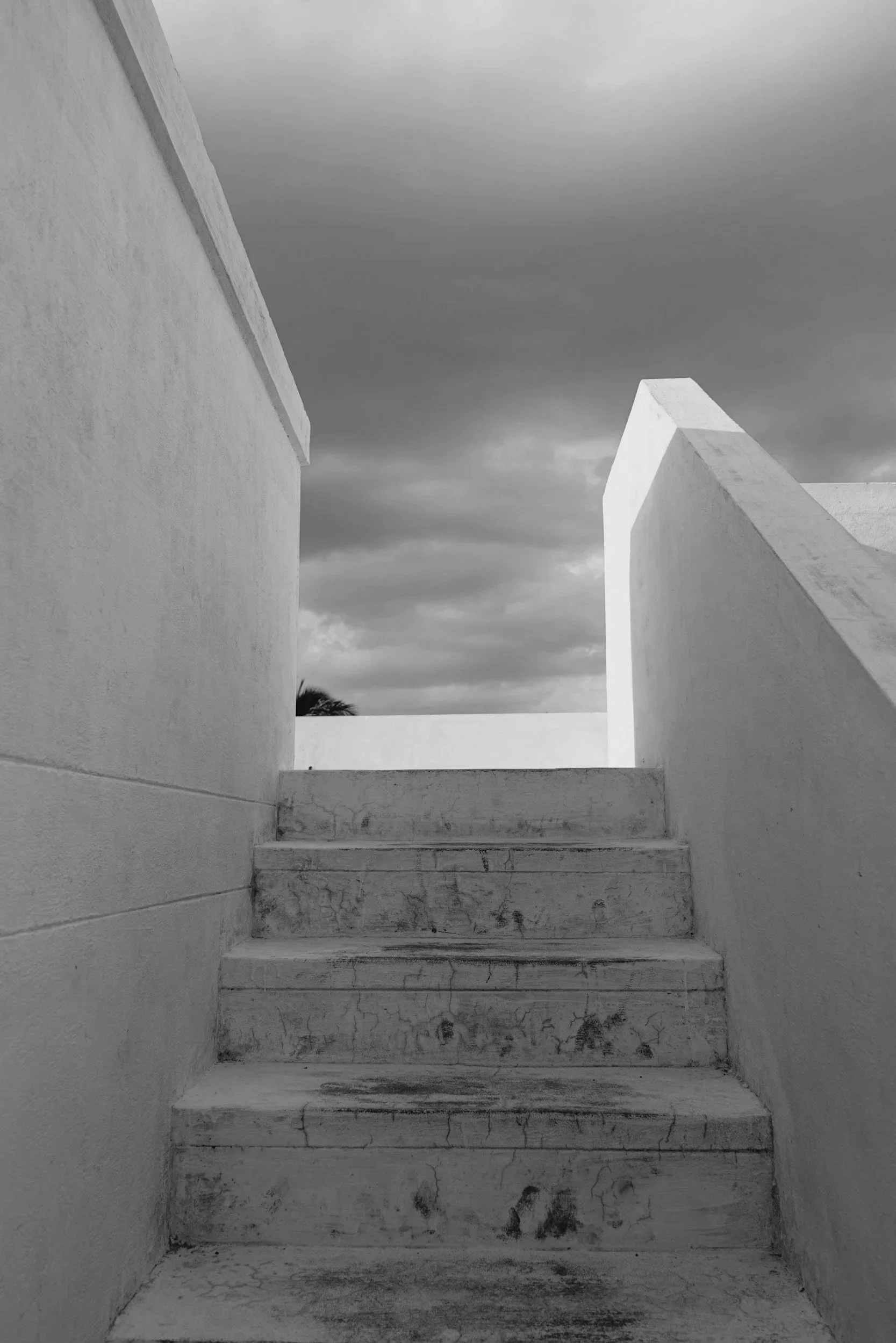 Black and white photo of concrete stairs leading up to an open space, with high white walls on either side and dark storm clouds in the sky above.