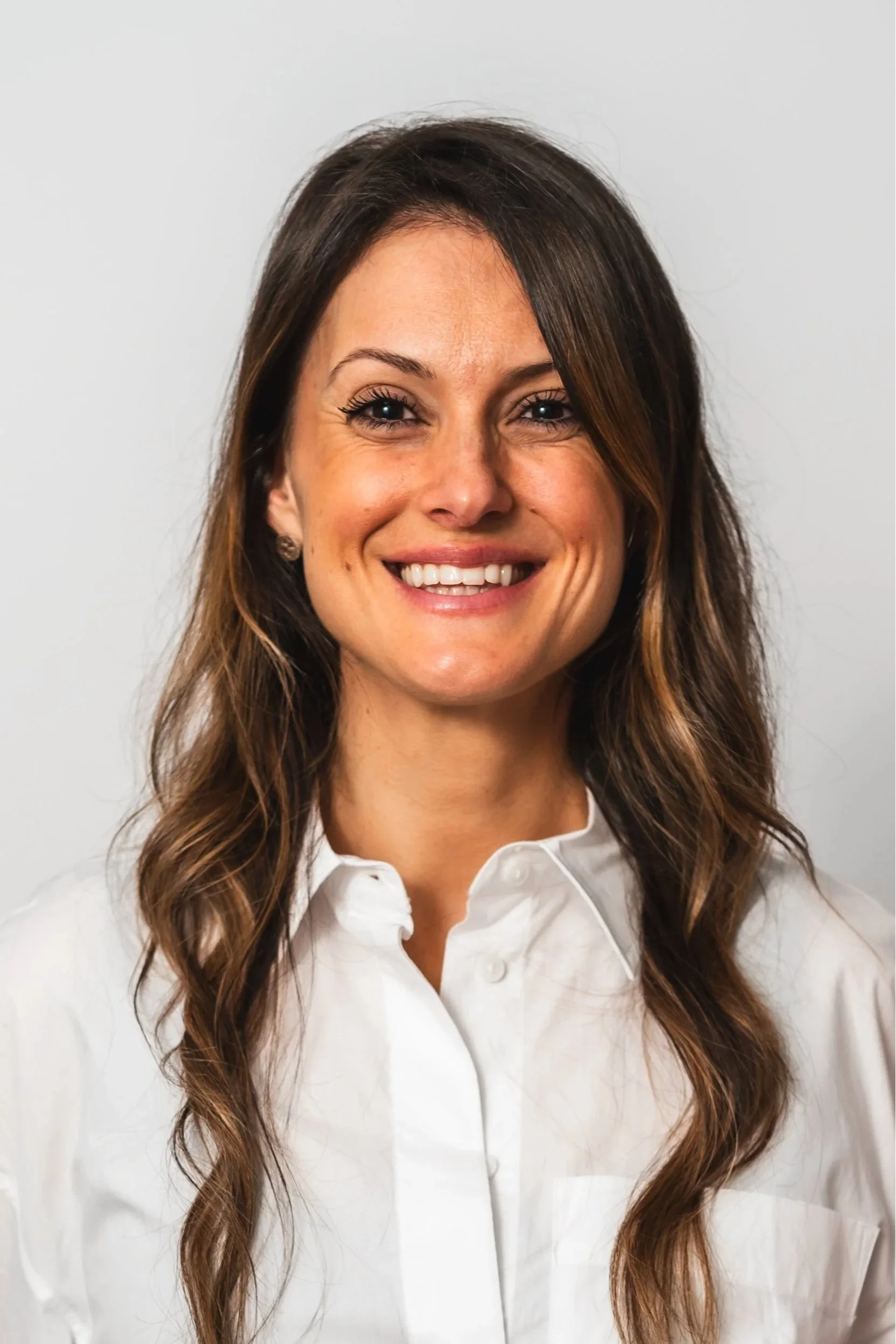Smiling woman with long brown hair wearing a white shirt against a plain background.