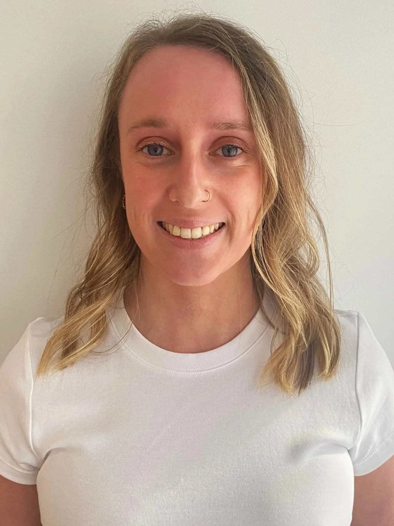 A woman with shoulder-length wavy blonde hair smiling, wearing a white T-shirt, standing against a plain white wall.