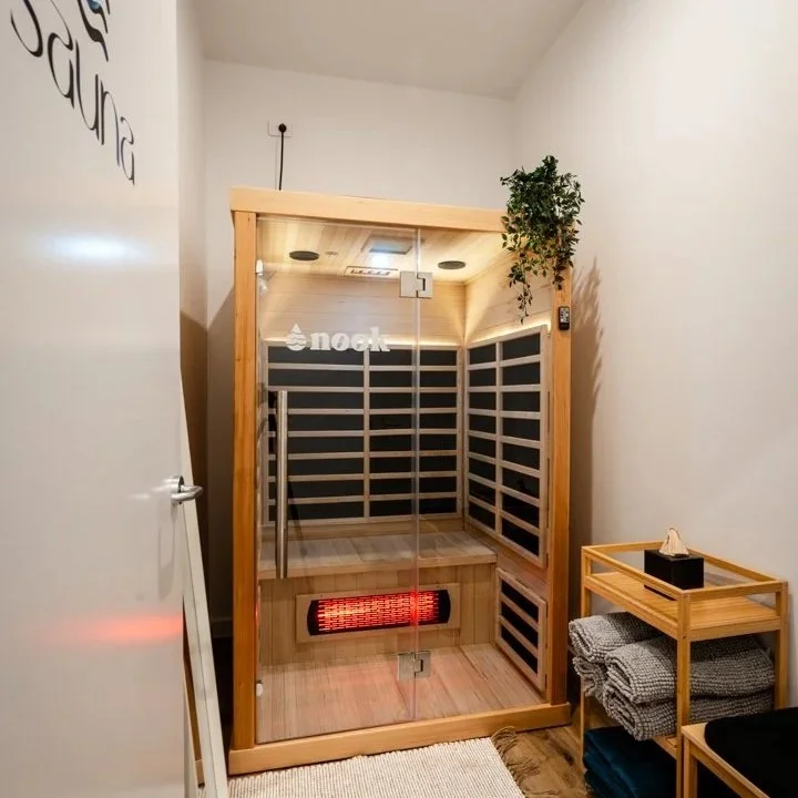 An infrared sauna with glass doors, wooden construction, and a hanging plant on top corner. Next to it is a small wooden table with folded towels and a tissue box.