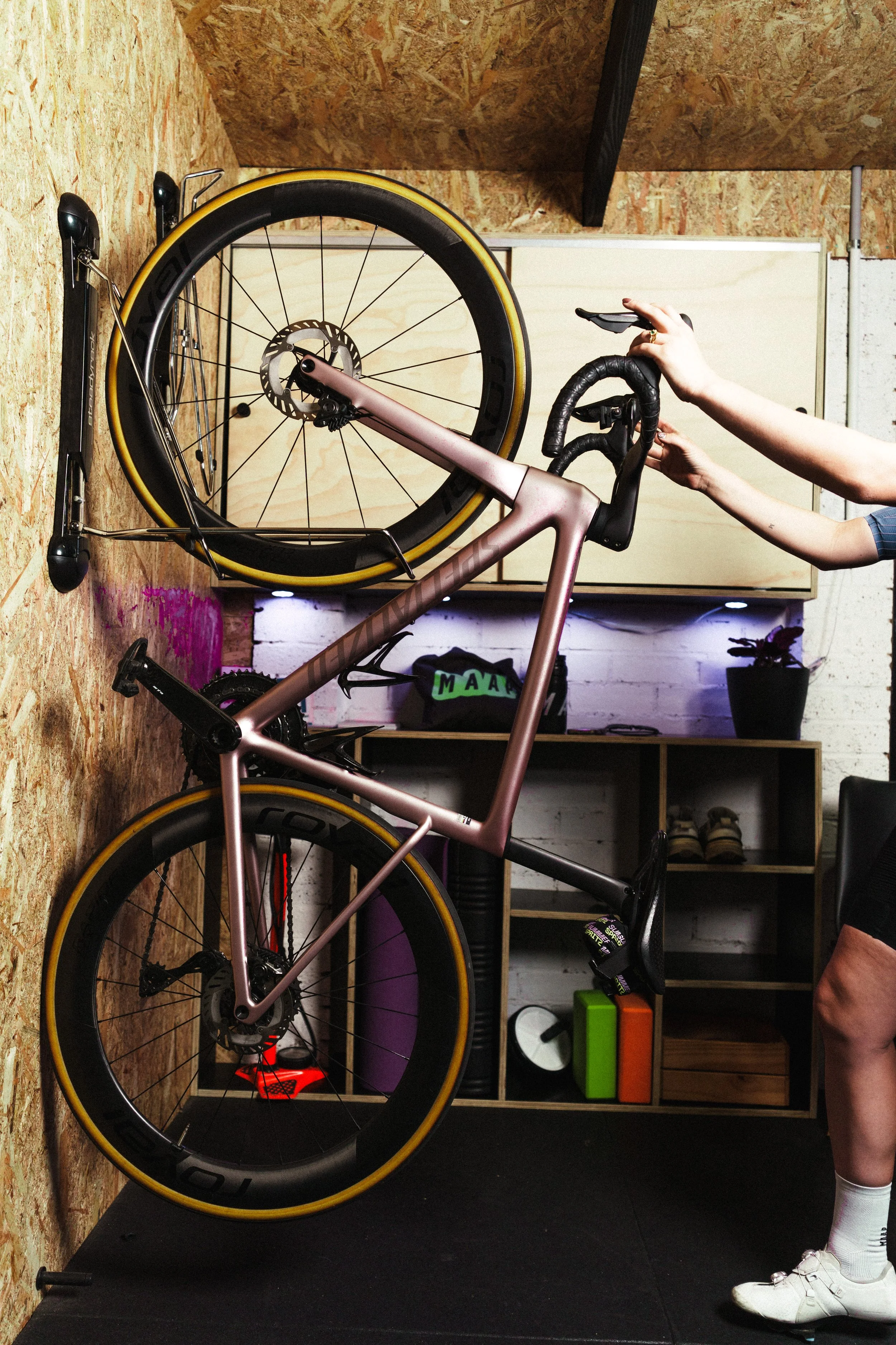A pink and black bicycle is mounted on a black rack against a wooden wall in a gym or workshop. A person wearing a blue shirt and white sneakers is adjusting the bike's handlebars. Shelves with fitness or gym equipment are visible in the background.