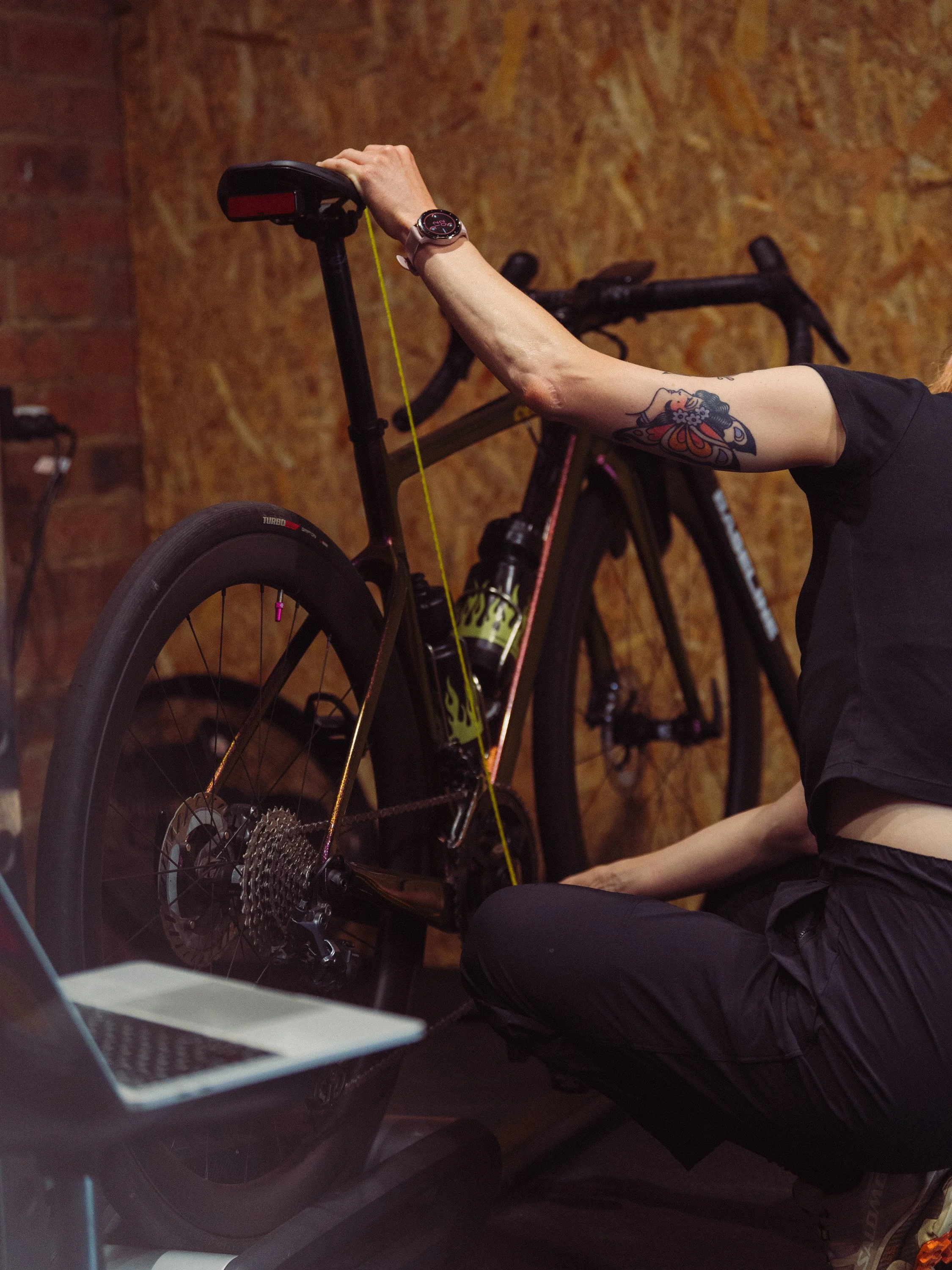 Person adjusting a black and green mountain bike, sitting on the floor, with a laptop nearby, against a wooden wall background.