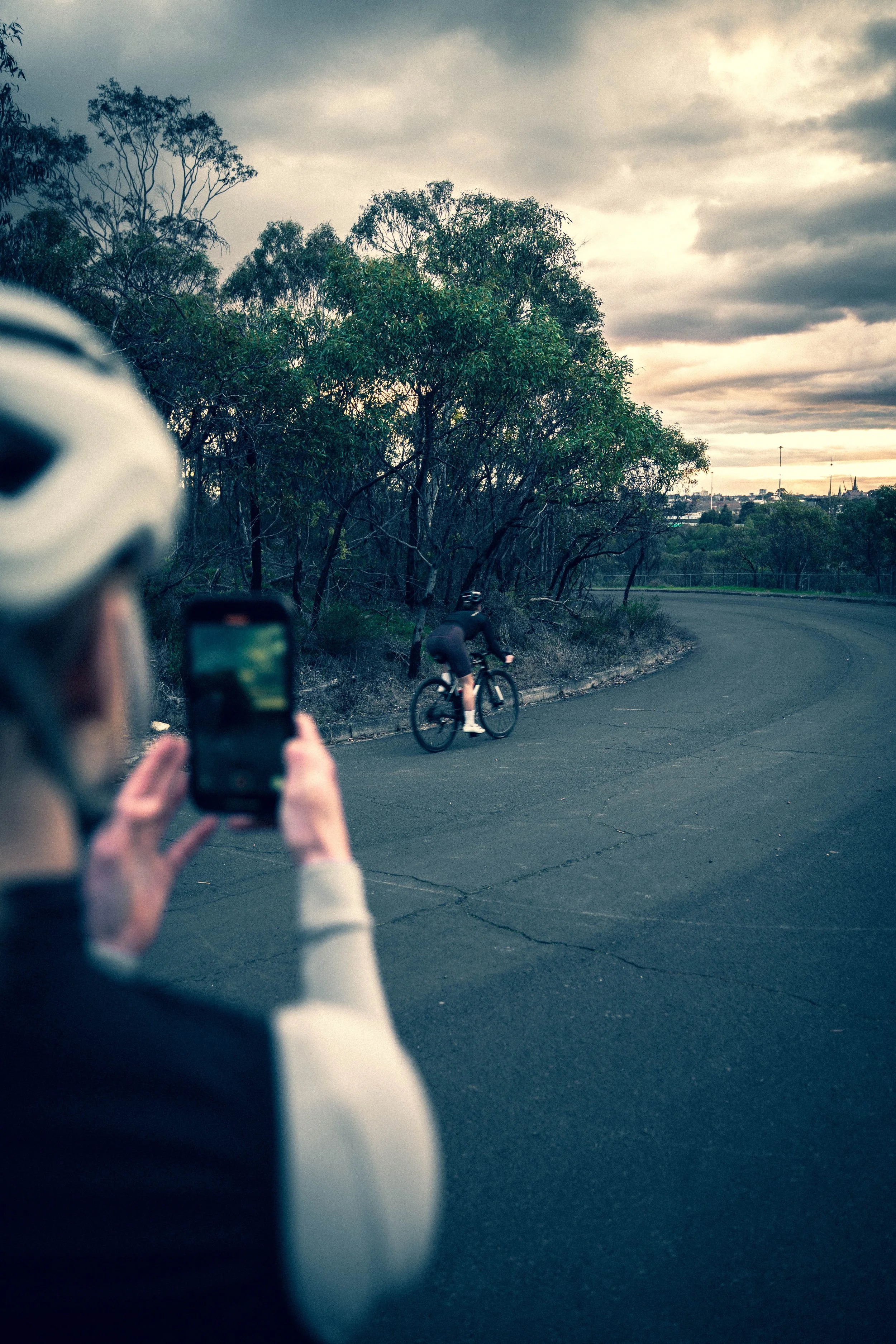 A person wearing a helmet is taking a photo of a cyclist riding along a curved road surrounded by trees, with a cloudy sky overhead.