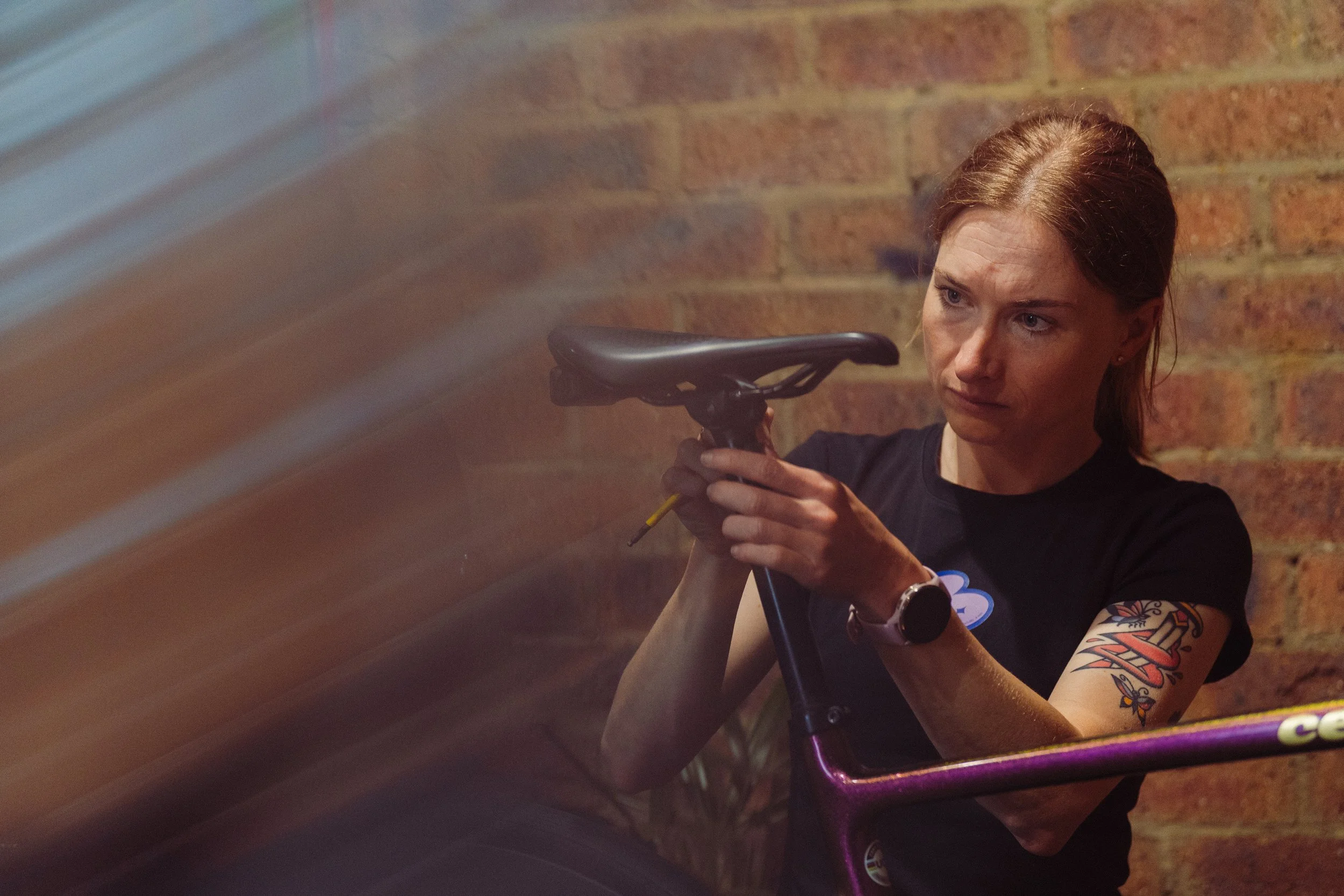 A woman with a tattoo on her arm and a watch on her wrist inspects or repairs a bicycle with a purple frame against a brick wall.