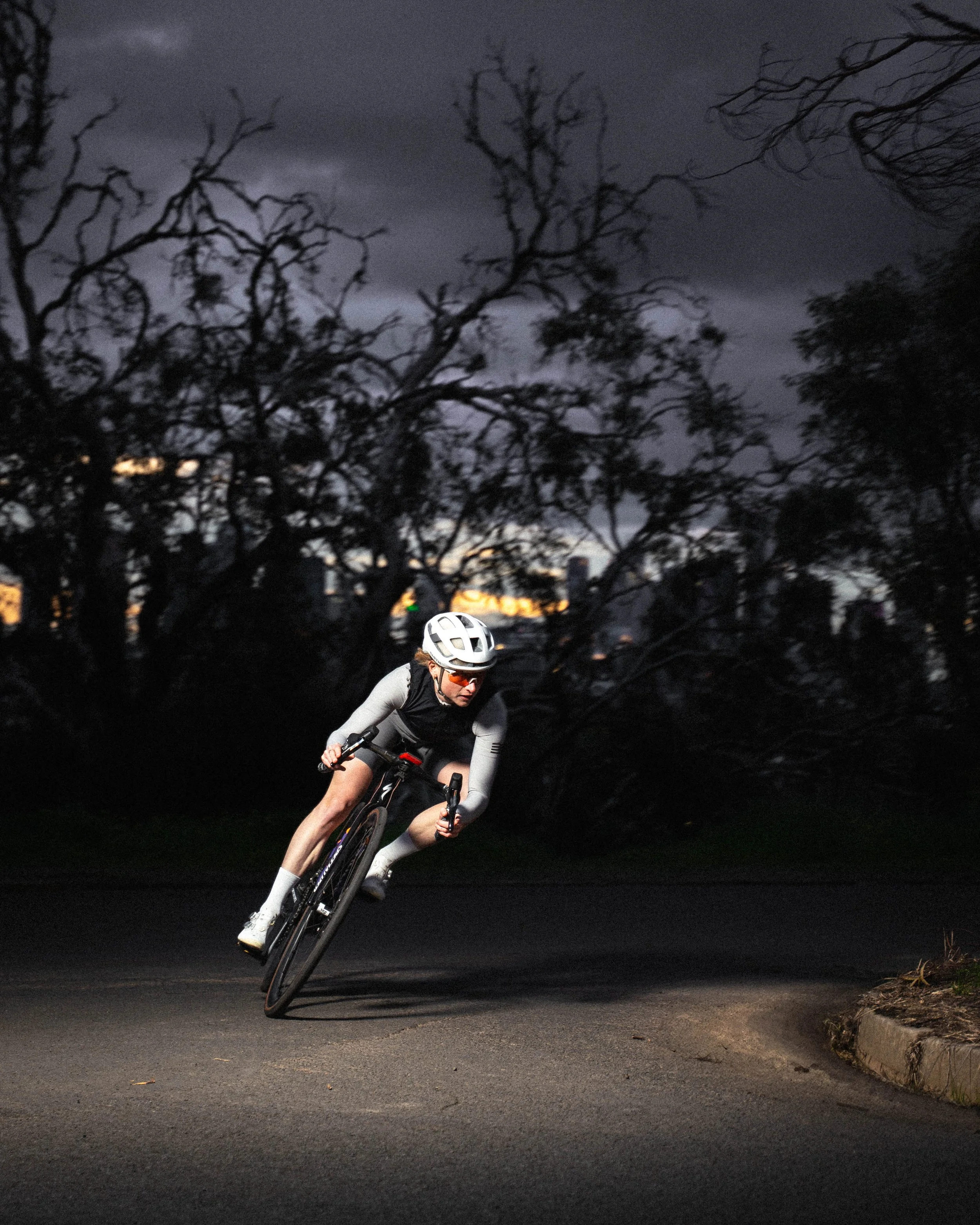 A cyclist wearing a white helmet and sunglasses riding a bike on a dark road during dusk, with leafless trees and a cloudy sky in the background.