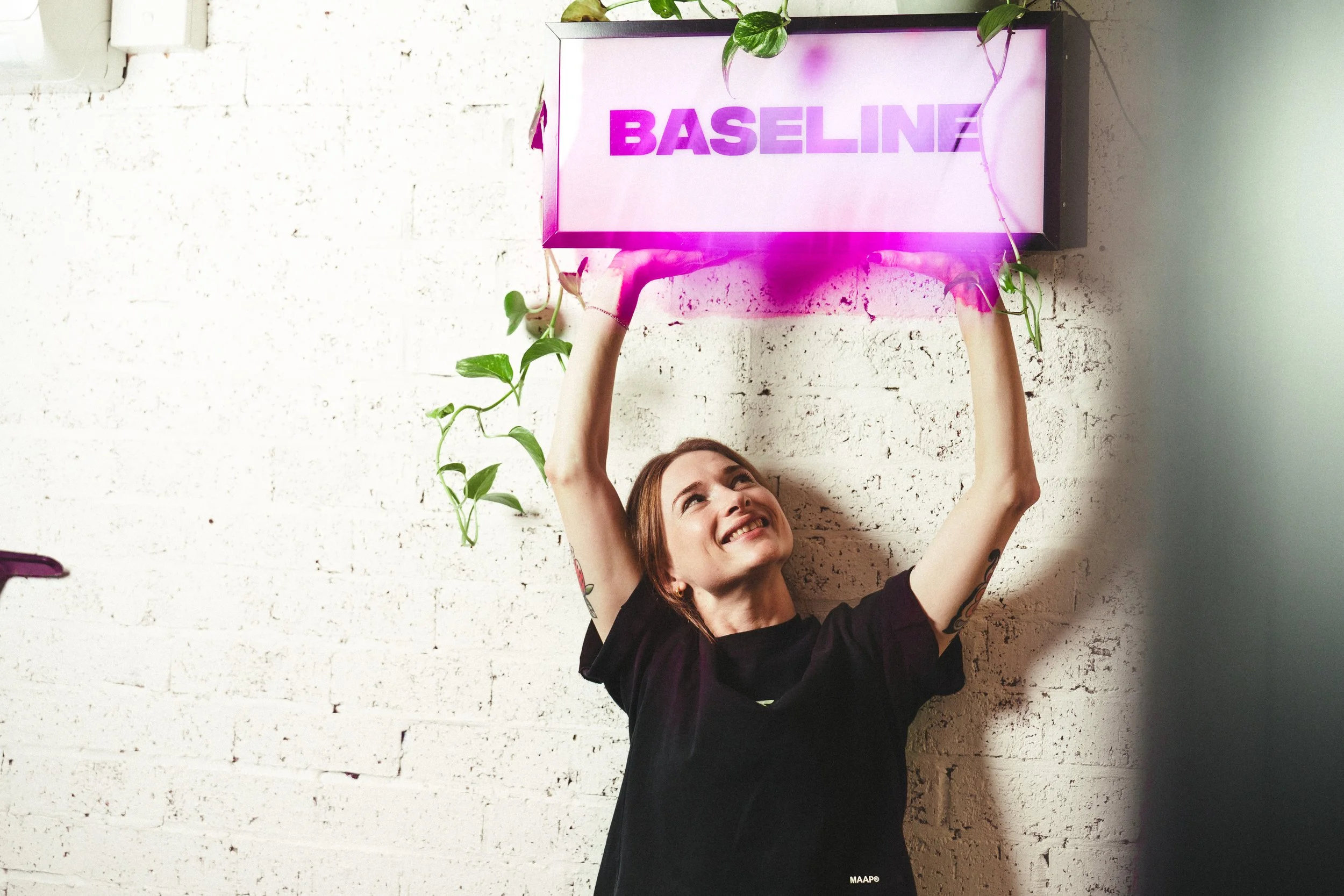 A woman holding a framed sign that says 'BASELINE' in purple letters, with a smile and arms raised, standing against a white brick wall decorated with green vines.