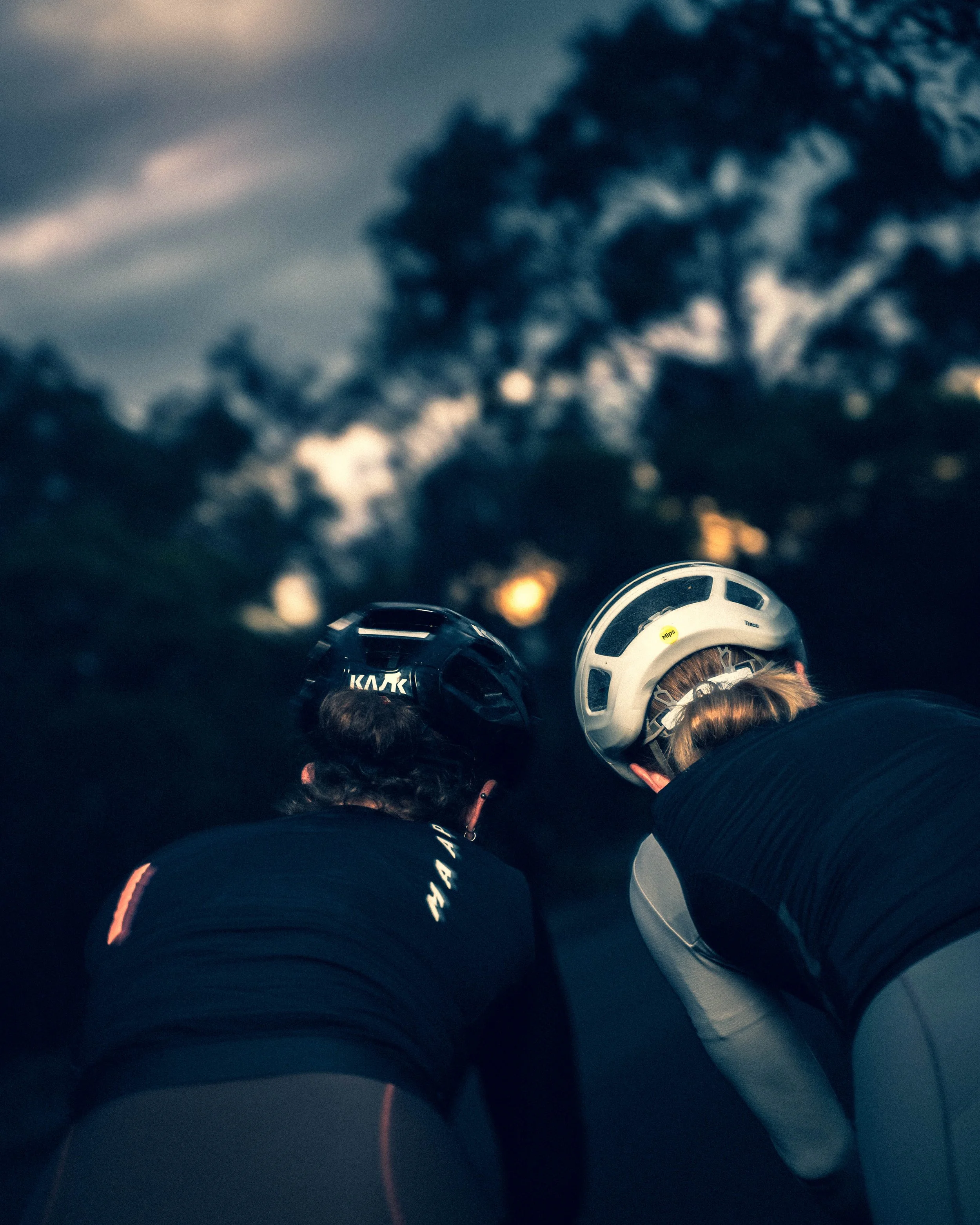 Two female cyclists in helmets and athletic clothing, one celebrating with a fist pump, helping fix a flat tire on a black and white road bike outdoors during dusk or dawn.