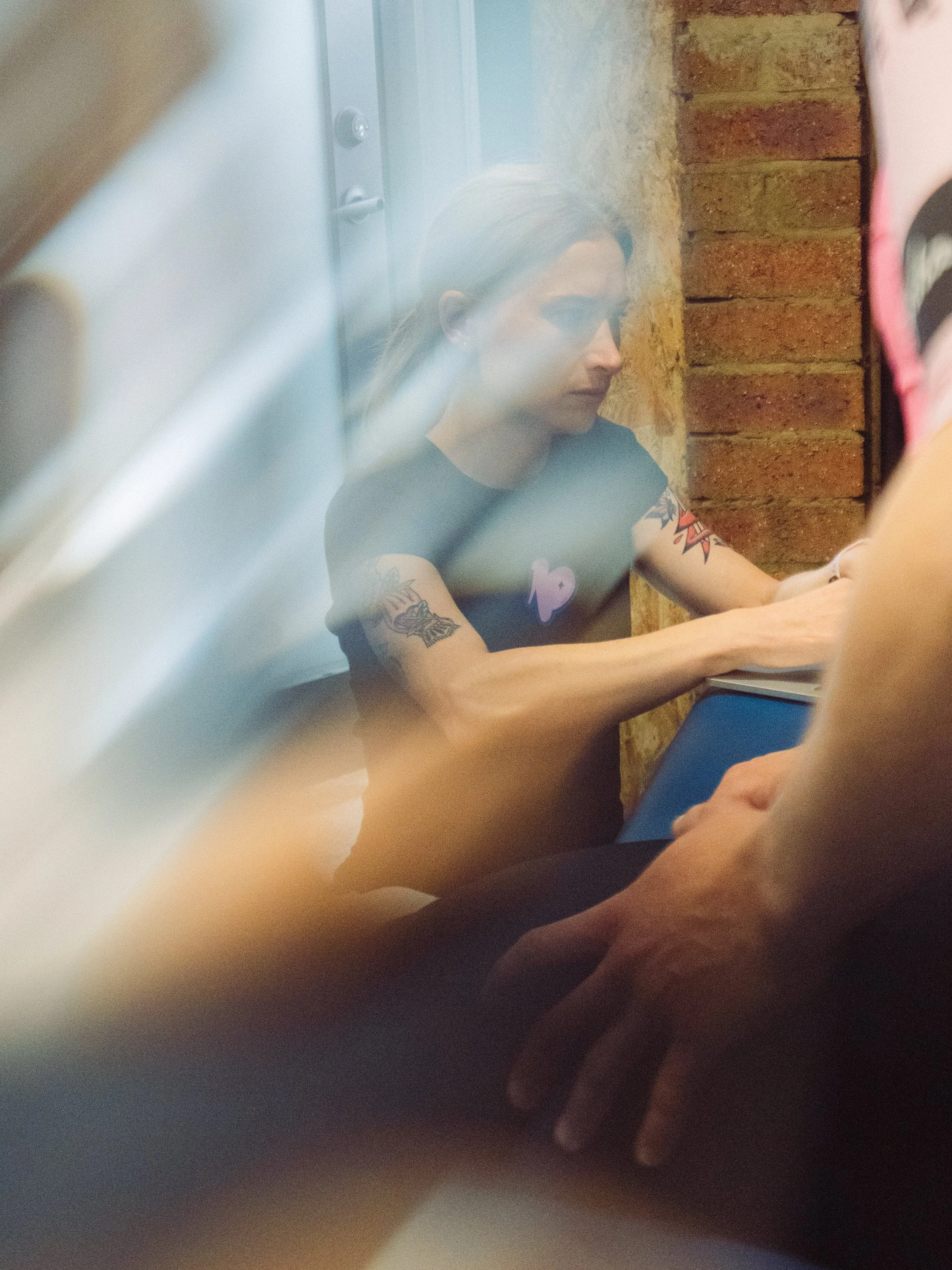 A woman with tattoos on her arms, sitting at a table with her hands resting on a laptop, seen through a foggy or reflective surface, with a brick wall in the background.