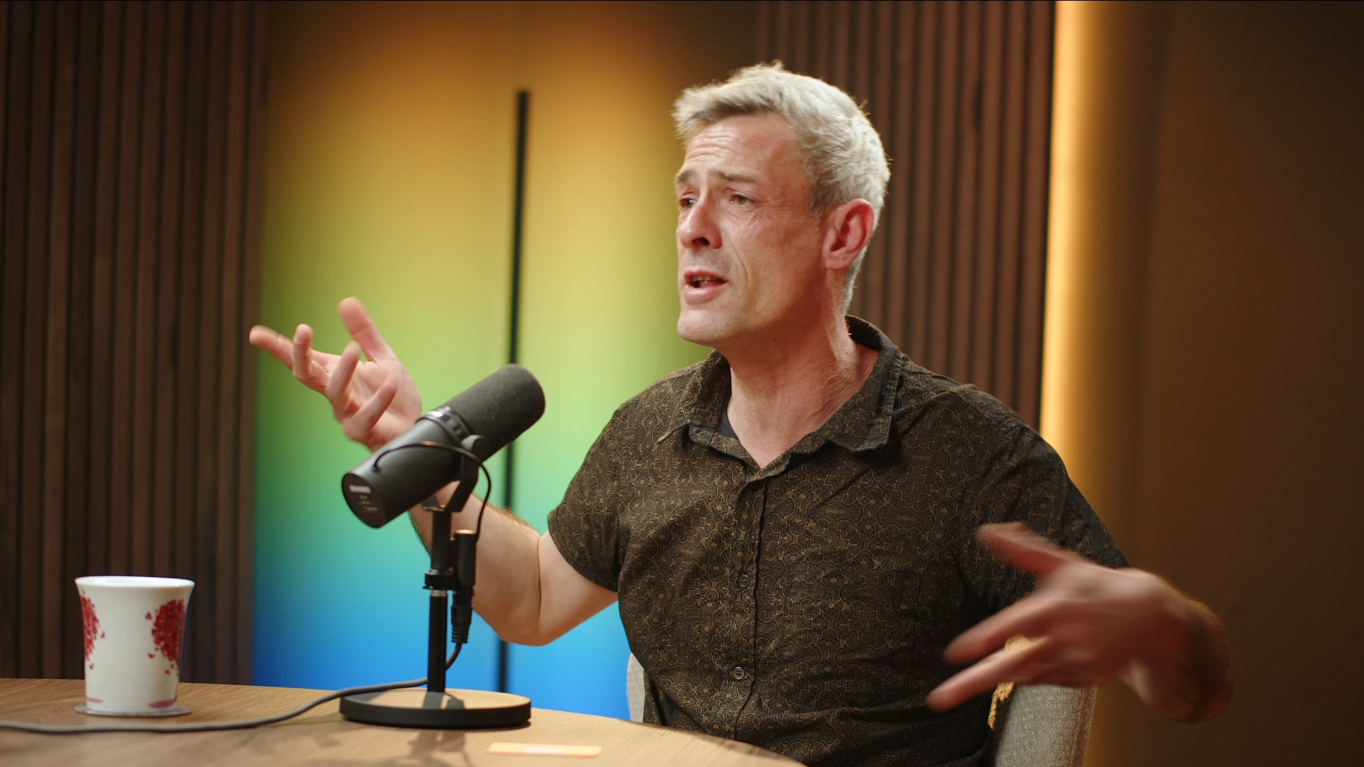 A middle-aged man with gray hair and a patterned dark shirt speaking into a microphone during a podcast or radio show, seated at a wooden table with a colorful blurred background.