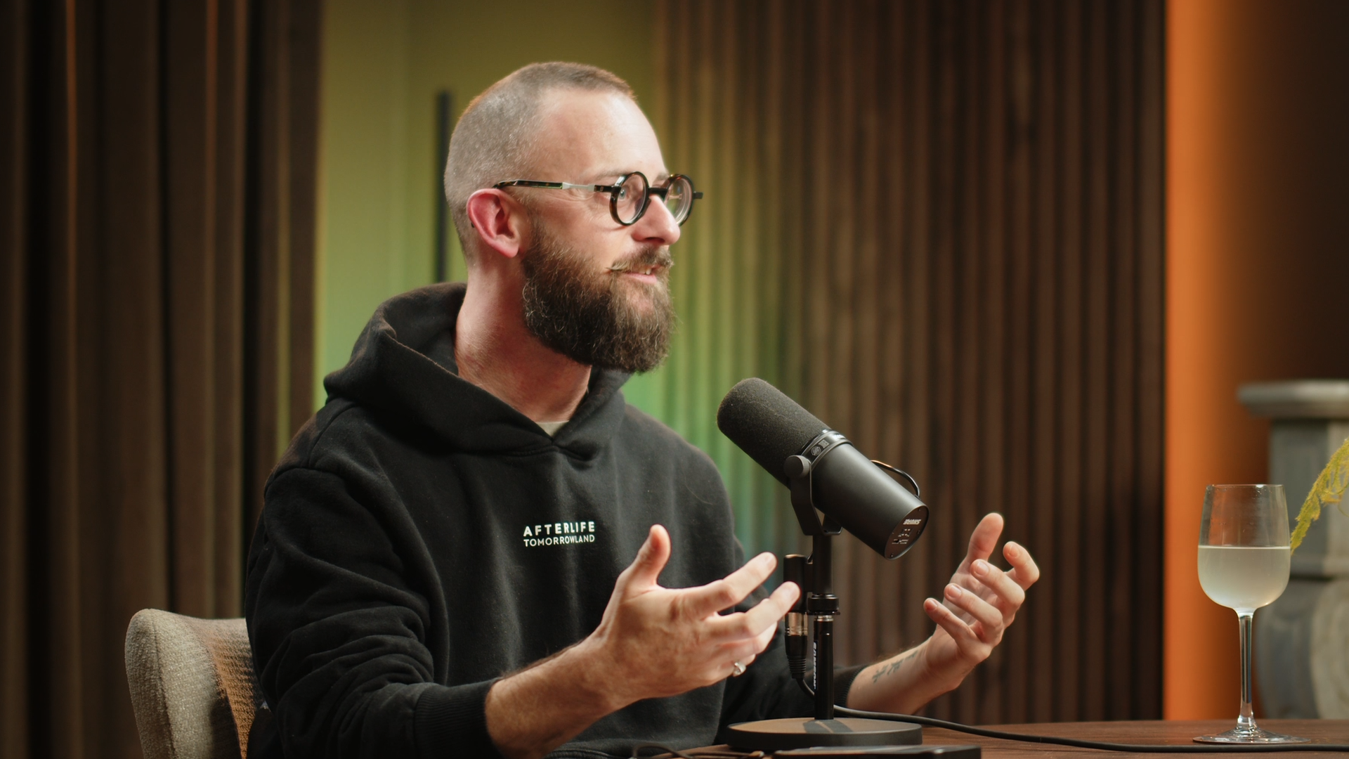 A man with glasses, a beard, and a shaved head wearing a black hoodie with the text 'AFTERLIFE TOMORROWLAND' sits at a table with a microphone, gesturing with his hands, in a room with dark curtains and warm lighting, and a glass of water on the table.