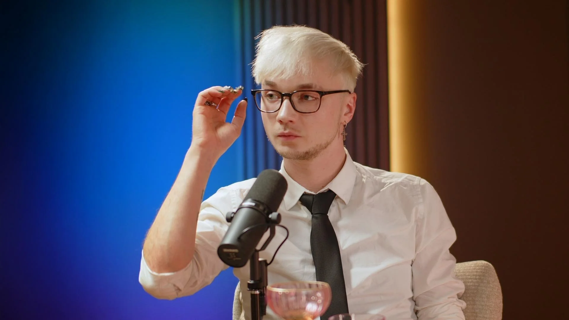 A young man with glasses, blonde hair, and earrings adjusting his glasses while sitting at a table with a microphone, a glass, and a pink bowl in front of him, in a brightly lit room with colorful curtains.
