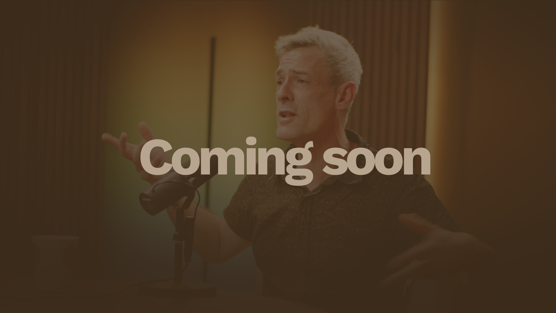 A middle-aged man with gray hair and a patterned dark shirt speaking into a microphone during a podcast or radio show, seated at a wooden table with a colorful blurred background.