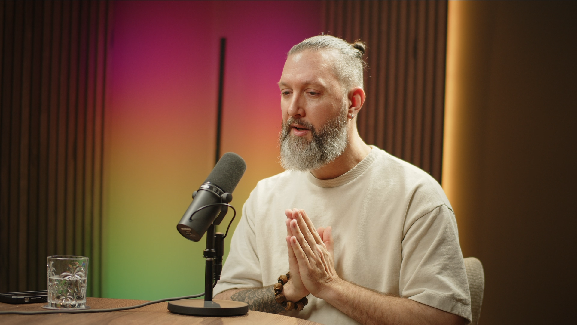 A bearded man with tattoos, wearing a beige t-shirt, seated at a table with a microphone, glass of water, and a laptop, appears to be speaking or praying with his hands together, in a room with colorful rainbow lighting.