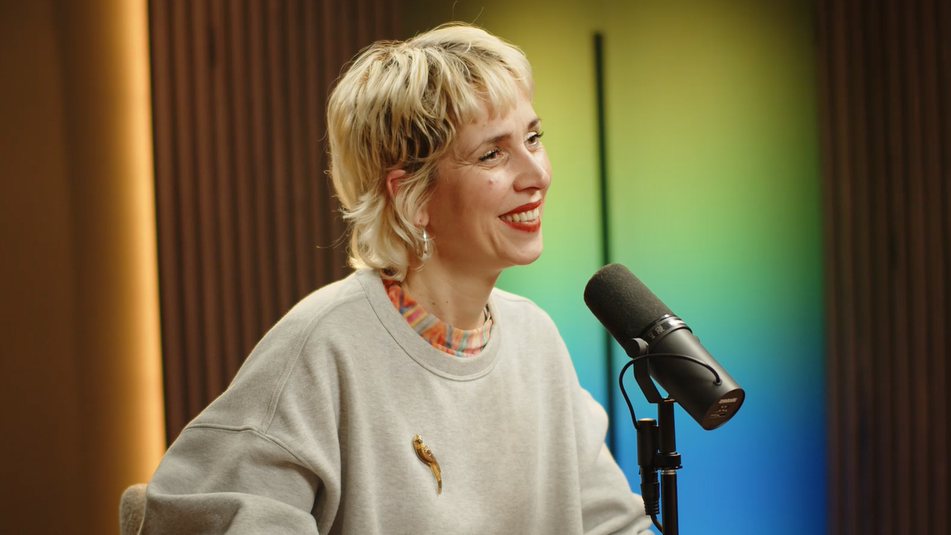 A woman with short blonde hair, wearing a gray sweatshirt and a colorful striped shirt underneath, sitting in front of a microphone, smiling.