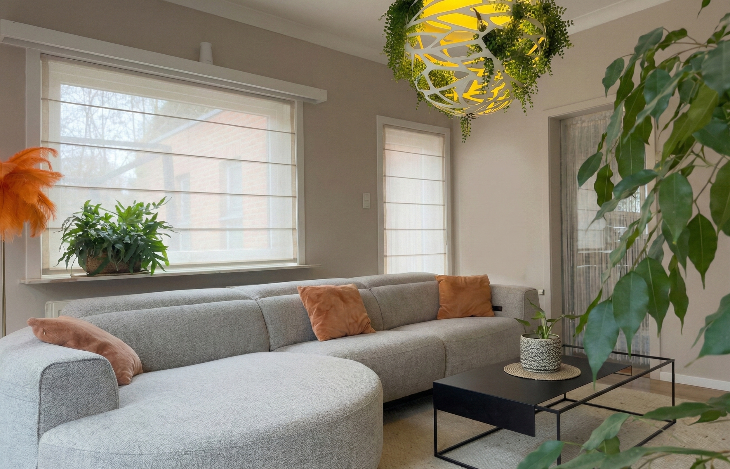Living room with a light gray sectional sofa with four orange pillows, a black coffee table with a potted plant, large windows with white blinds, and a decorative green and yellow hanging light fixture.