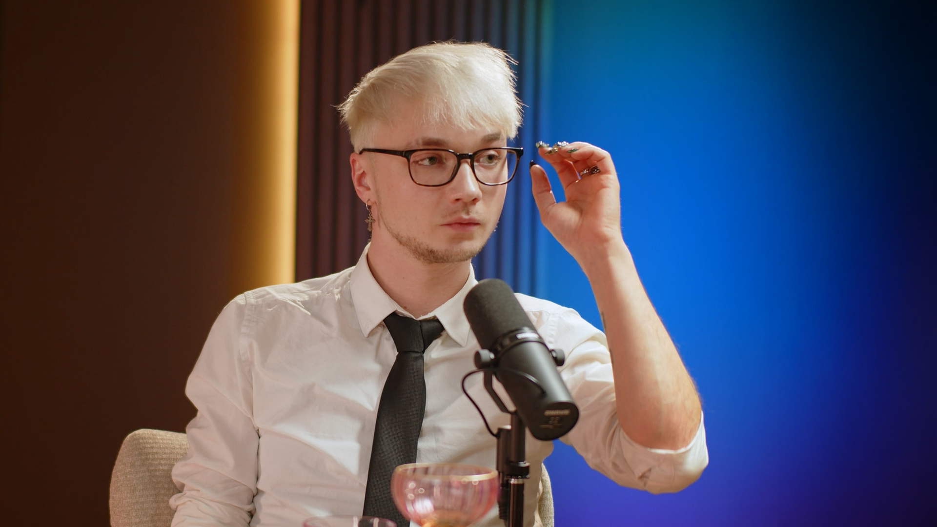 A young man with blonde hair, glasses, and earrings, wearing a white shirt and black tie, is adjusting glasses while sitting in front of a microphone, with a colorful background.