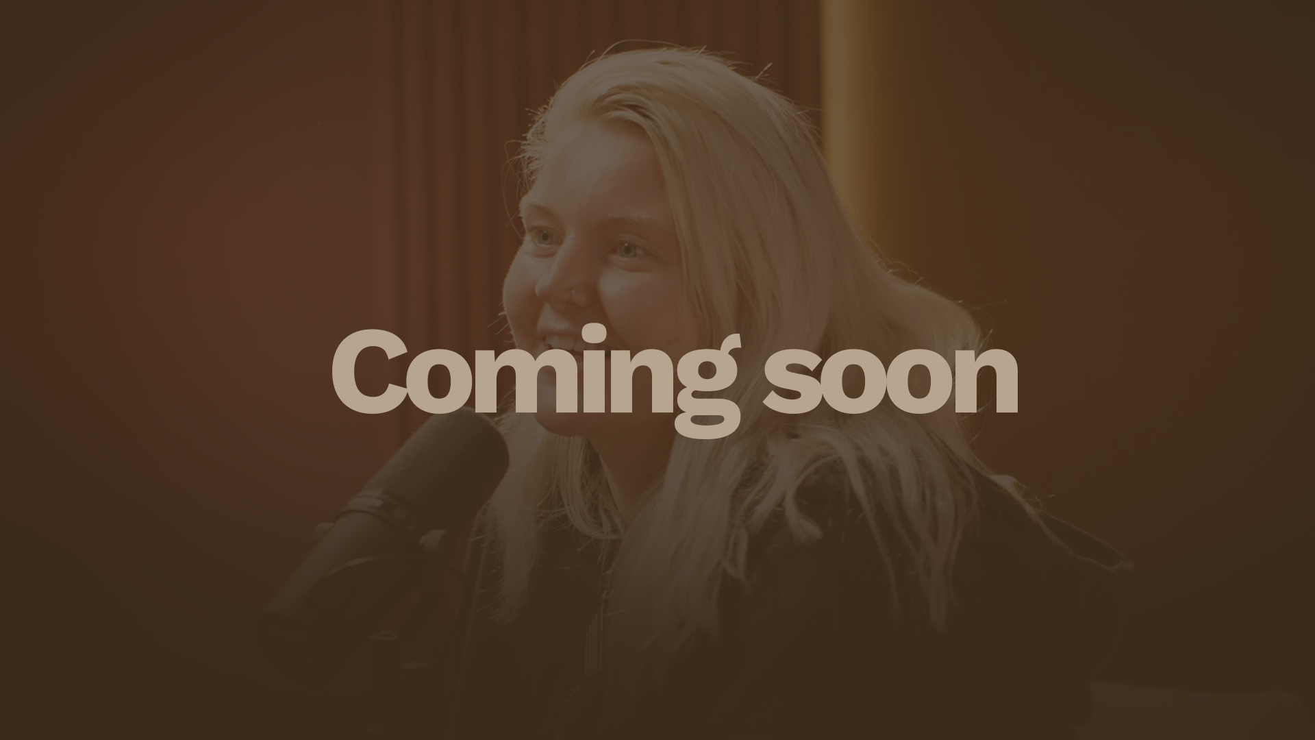 A woman with long blonde hair and a nose ring speaking into a microphone in a recording studio.