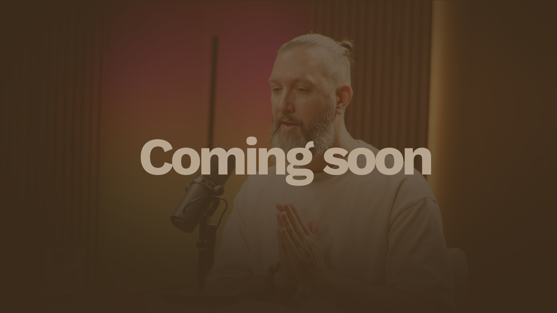 A man with a beard, styled hair, wearing a light-colored t-shirt, sitting at a table with a microphone, a glass of water, and a laptop, in a studio with colorful gradient background.