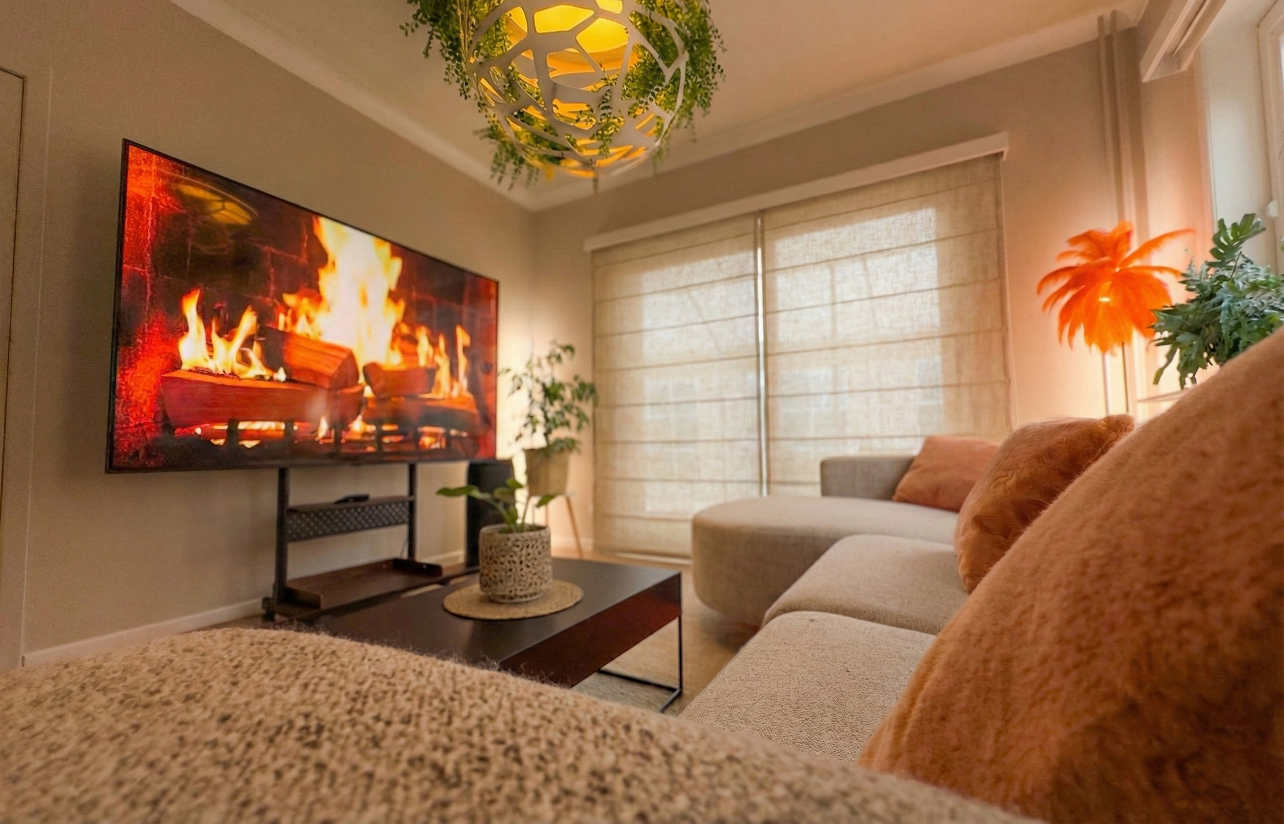 Cozy living room with a large TV displaying a fireplace, beige sofa with orange cushions, plants, and window with blinds.
