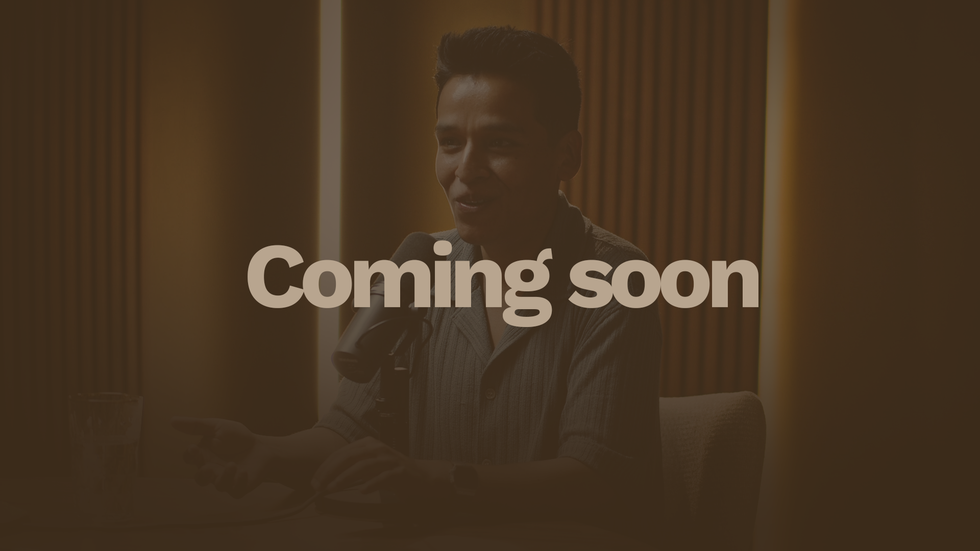 A smiling man with black hair and a blue striped shirt is sitting at a table in front of a microphone during a recording or interview session. There is a glass of water on the table and a modern, wooden-paneled wall in the background.