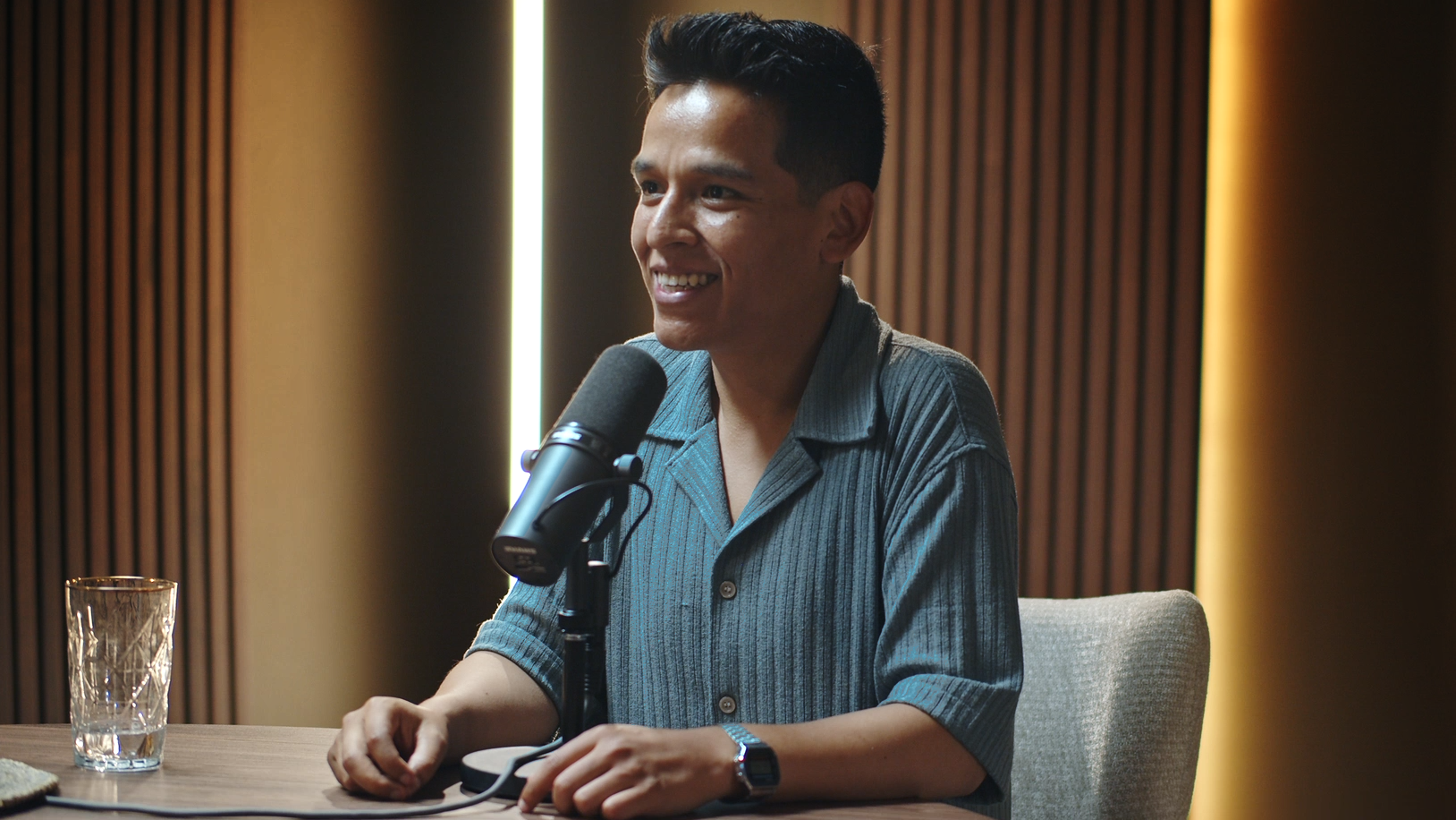 A smiling man with black hair and a blue striped shirt is sitting at a table in front of a microphone during a recording or interview session. There is a glass of water on the table and a modern, wooden-paneled wall in the background.
