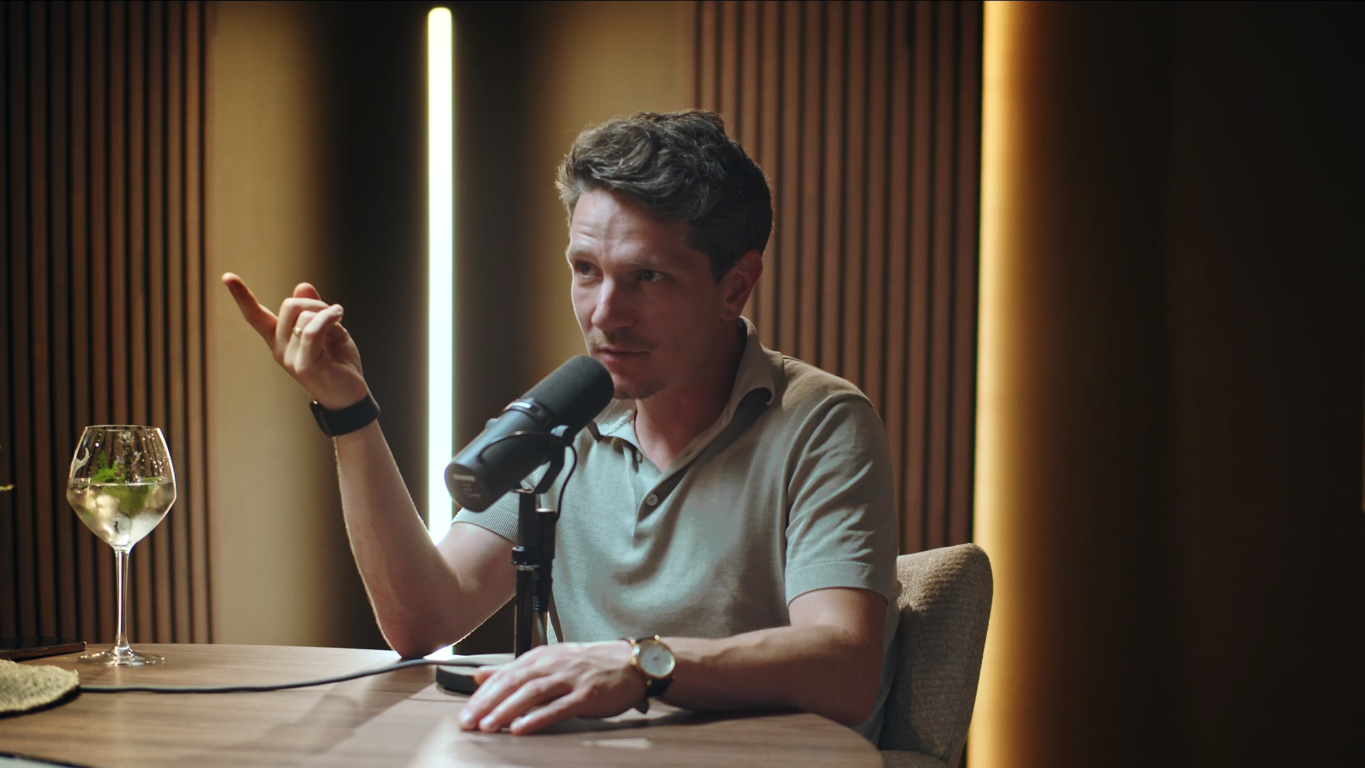 A man seated at a table with a microphone, gesturing with his hand, and a glass of sparkling water with mint leaves on the table.
